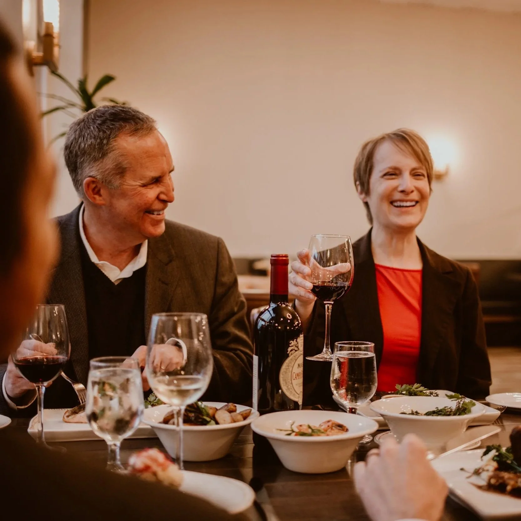 A middle-aged man and woman are sitting at a table at Maxwell's Steak & Seafood, smiling and enjoying a meal with wine. The woman is holding a glass of red wine. Plates of food and glasses of water are also visible on the table.