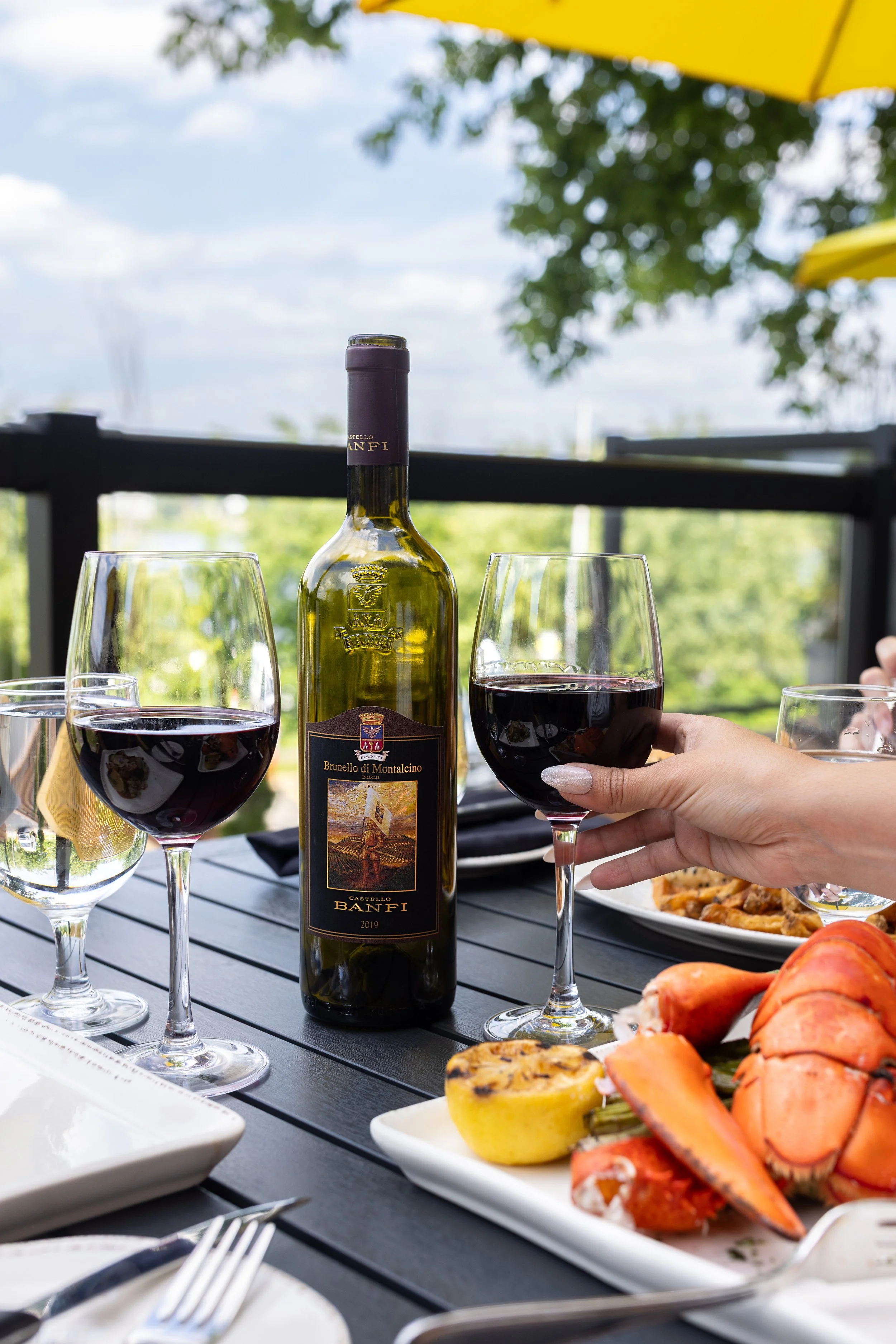 A table on Maxwell's Steak & Seafood's patio with a bottle of red wine, three glasses of red wine, a plate of lobster, a lemon, and a plate with some food, with a yellow umbrella overhead and trees in the background.