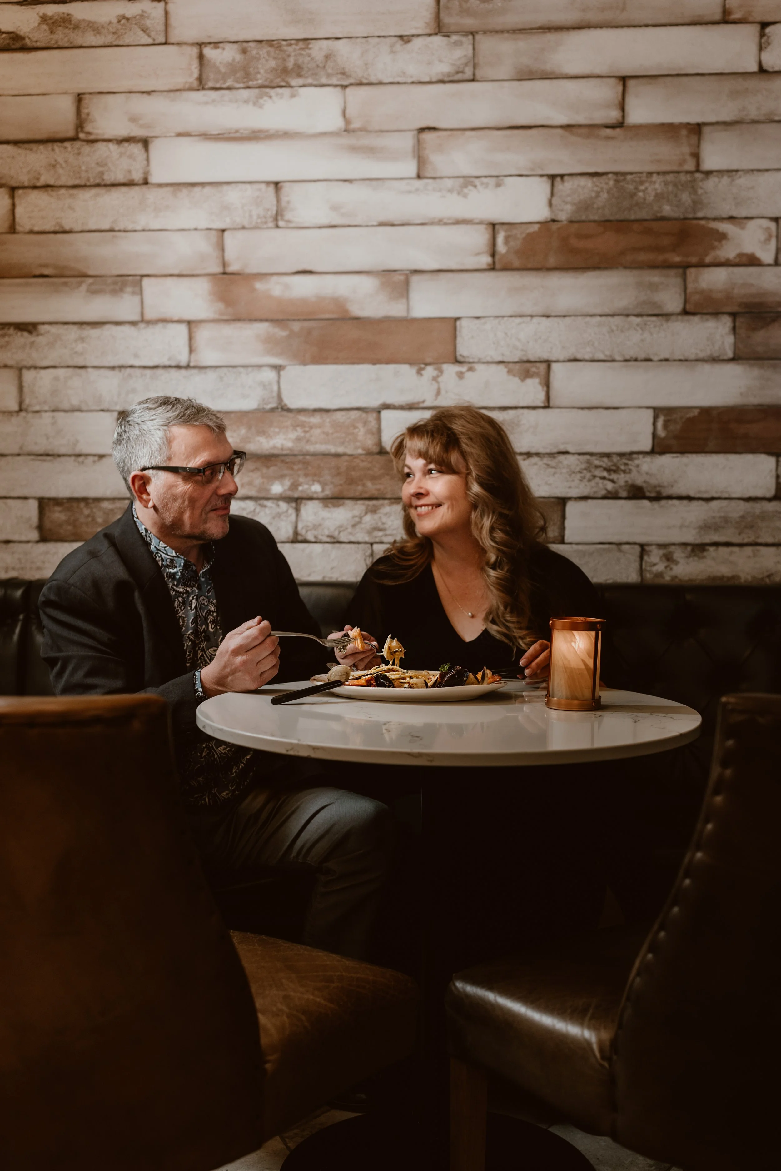 A man and woman sitting at a table in Maxwell's Steak & Seafood lounge, smiling at each other while sharing a delicious meal, with a brick wall background and a candle on the table.