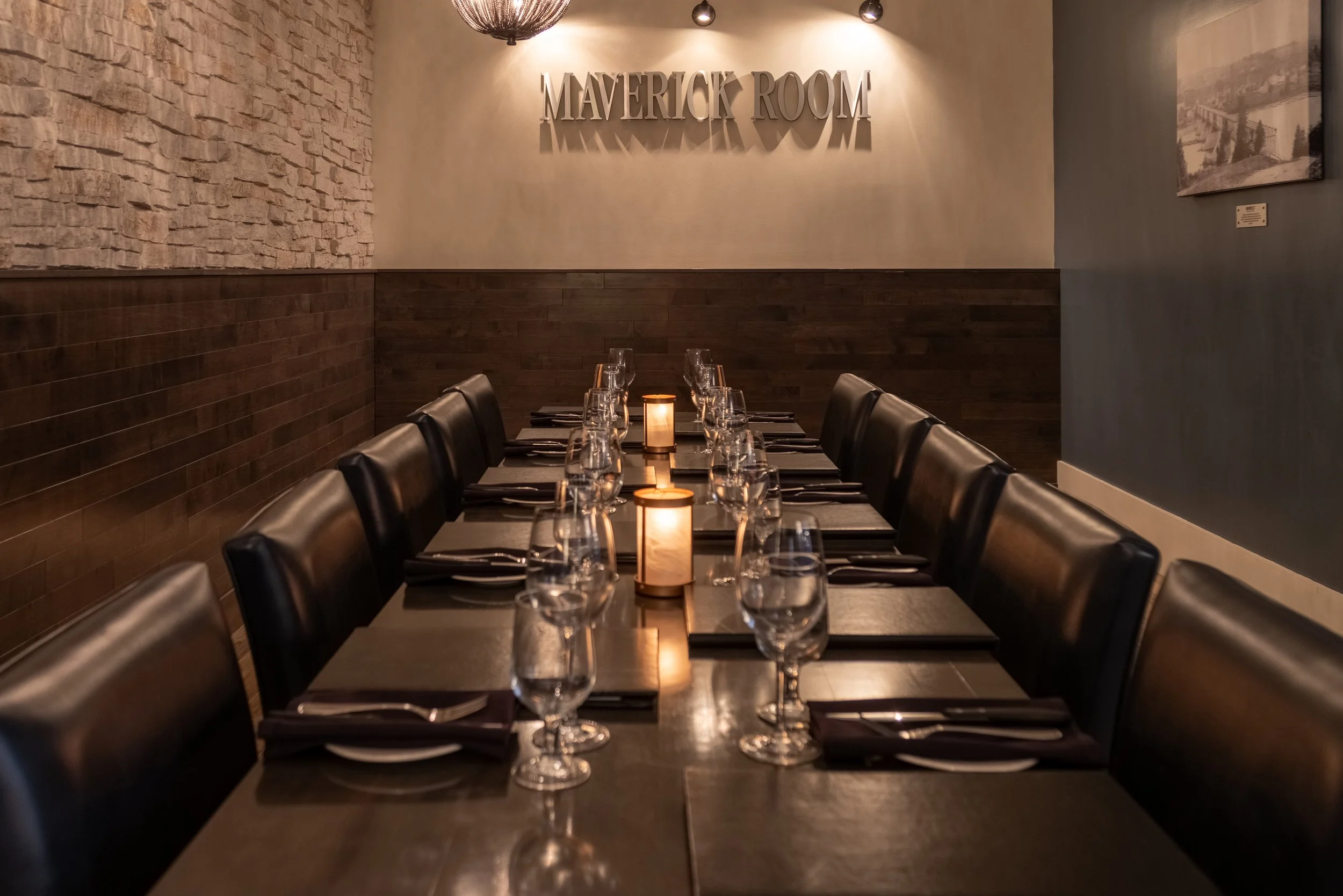 The private Maverick room at Maxwell's with a long table set for a meal, featuring black chairs, wine glasses, and black napkins, illuminated by a candle and overhead lighting, with a sign that reads 'MAVERICK ROOM' on the wall.