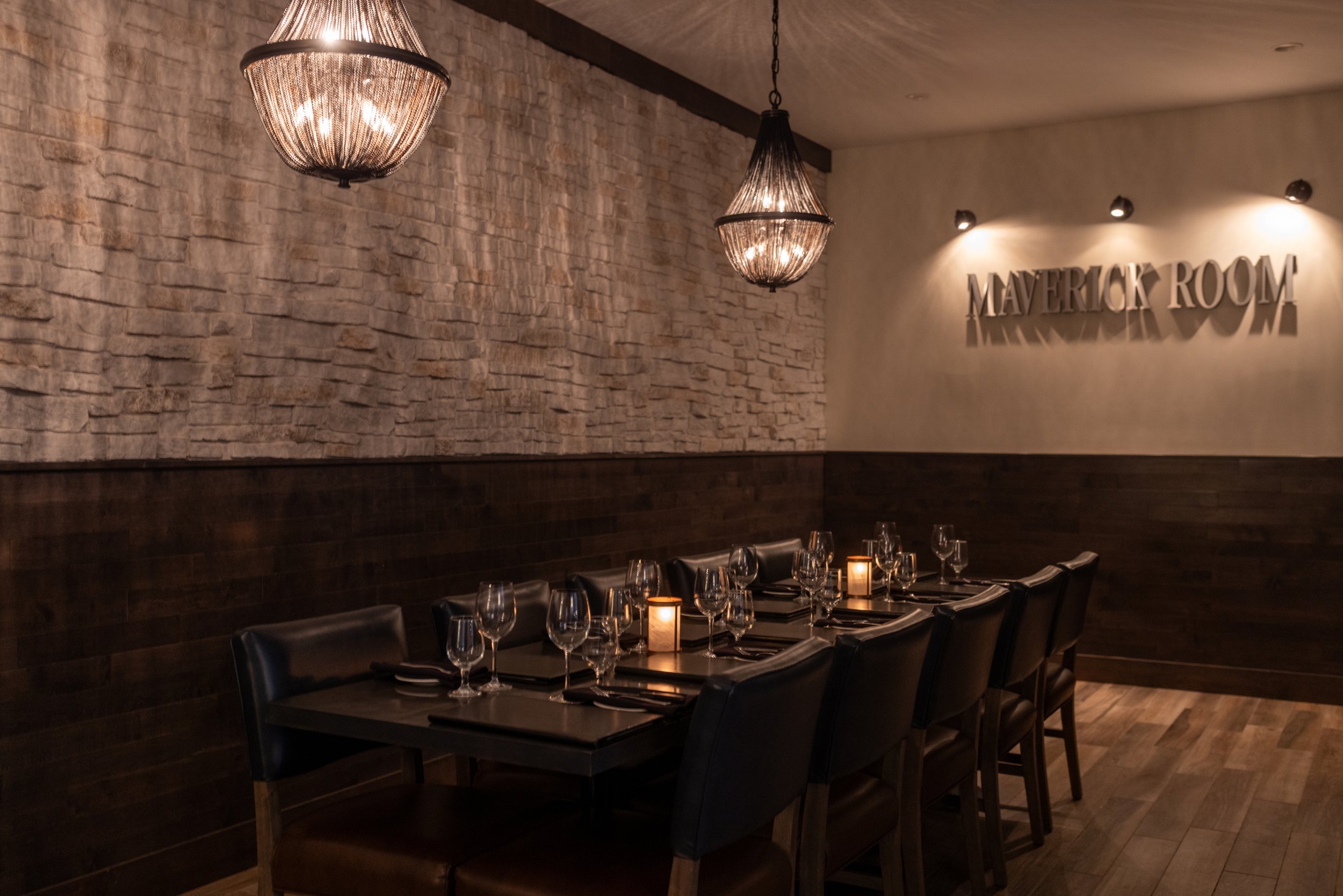 The dimly lit Maverick room's table set with wine glasses and candles against a brick and painted wall, with a sign reading 'Maverick Room' on the wall inside Maxwell's Steak & Seafood.