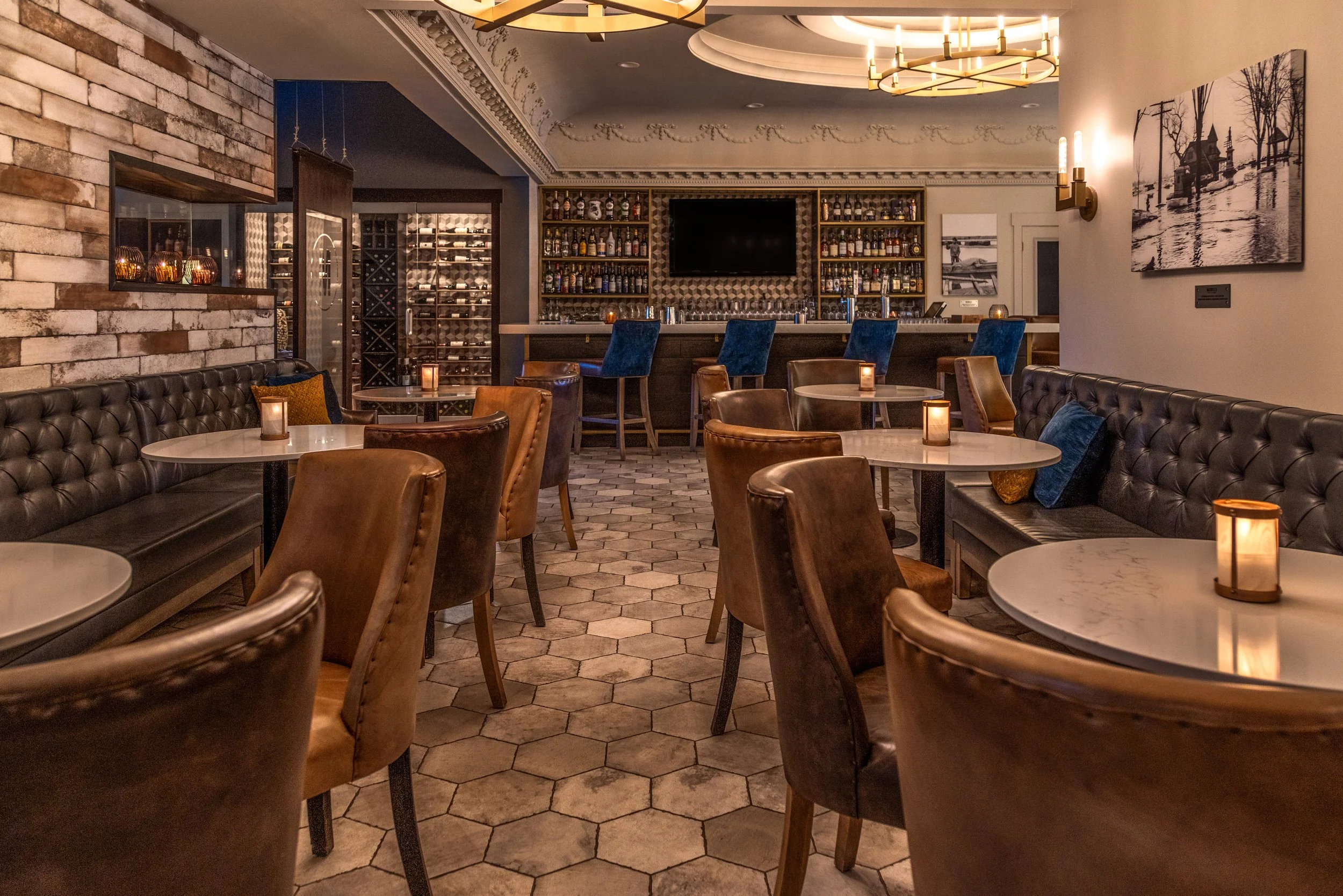 The cozy inside of Maxwell's  bar with hexagonal tiled floor, leather and fabric chairs around marble-topped tables, soft candle lighting, a brick accent wall, a bar with a variety of liquor bottles, wall art, and ambient ceiling lights.