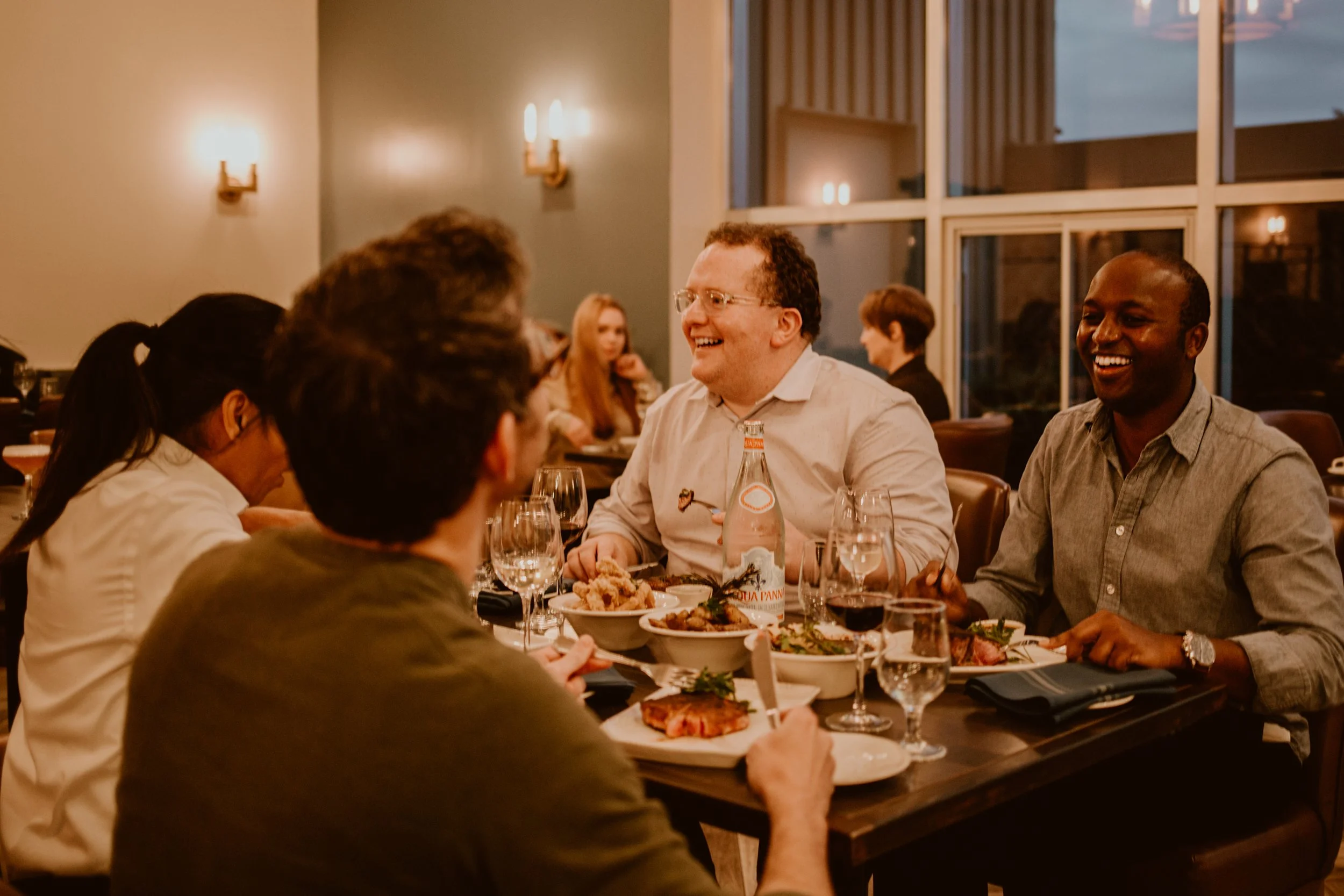 People sitting at a restaurant table in Maxwell's Steak & Seafood enjoying a conversation and meal, with smiles and glasses of wine, in a warmly lit setting.