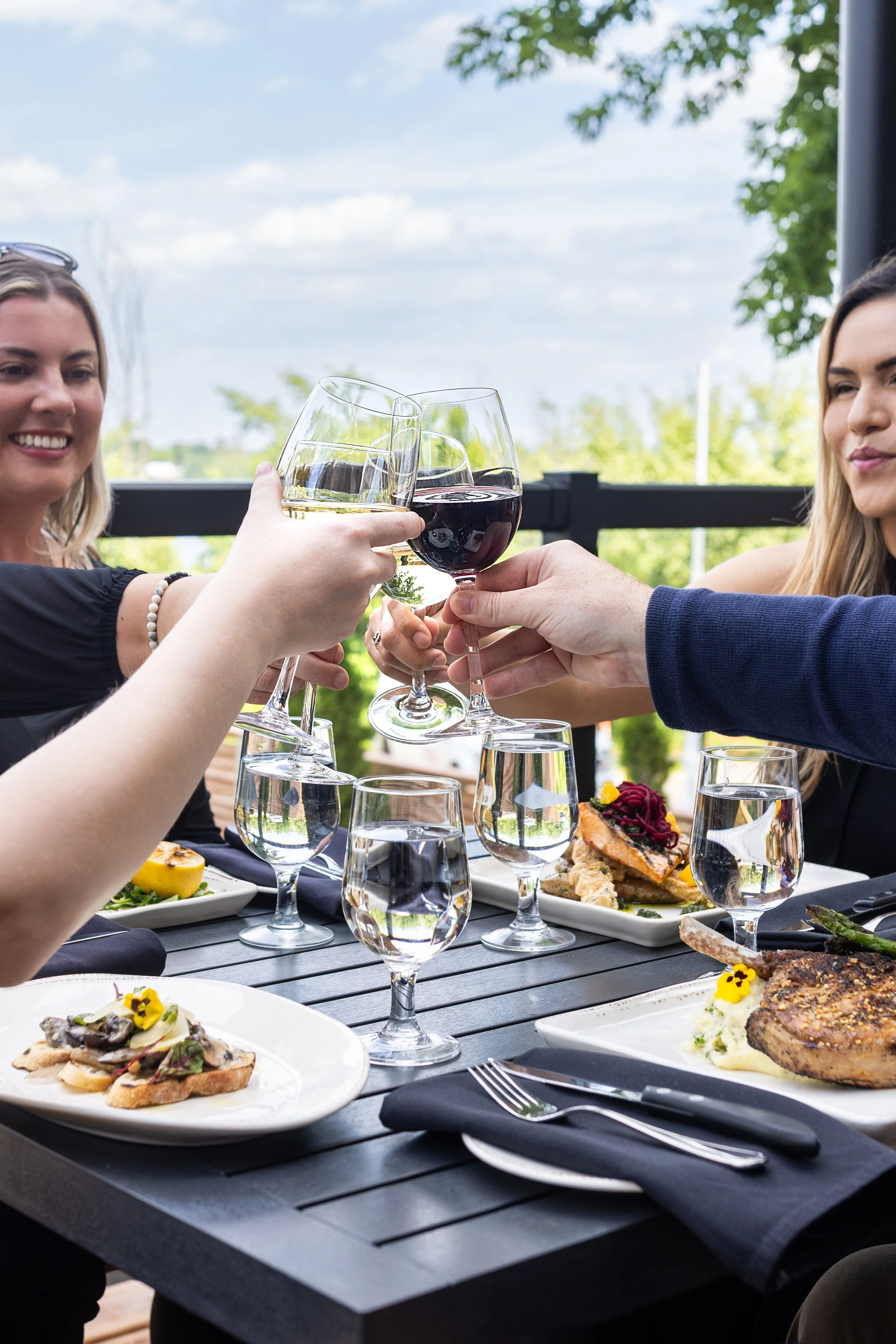 People clinking wine glasses during a meal the patio of Maxwell's Steak & Seafood, with plates of food including steak, and a scenic outdoor background with trees.
