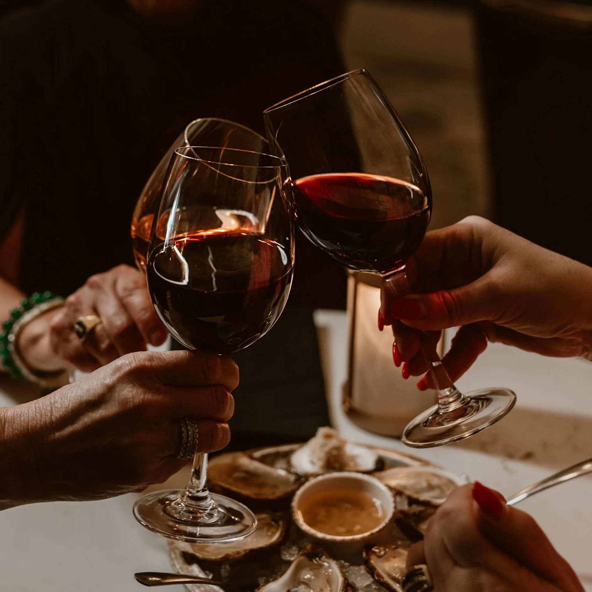 Three people clink glasses of red wine over a table of seafood, including oysters and a dipping sauce at Maxwell's Steak & Seafood restaurant.
