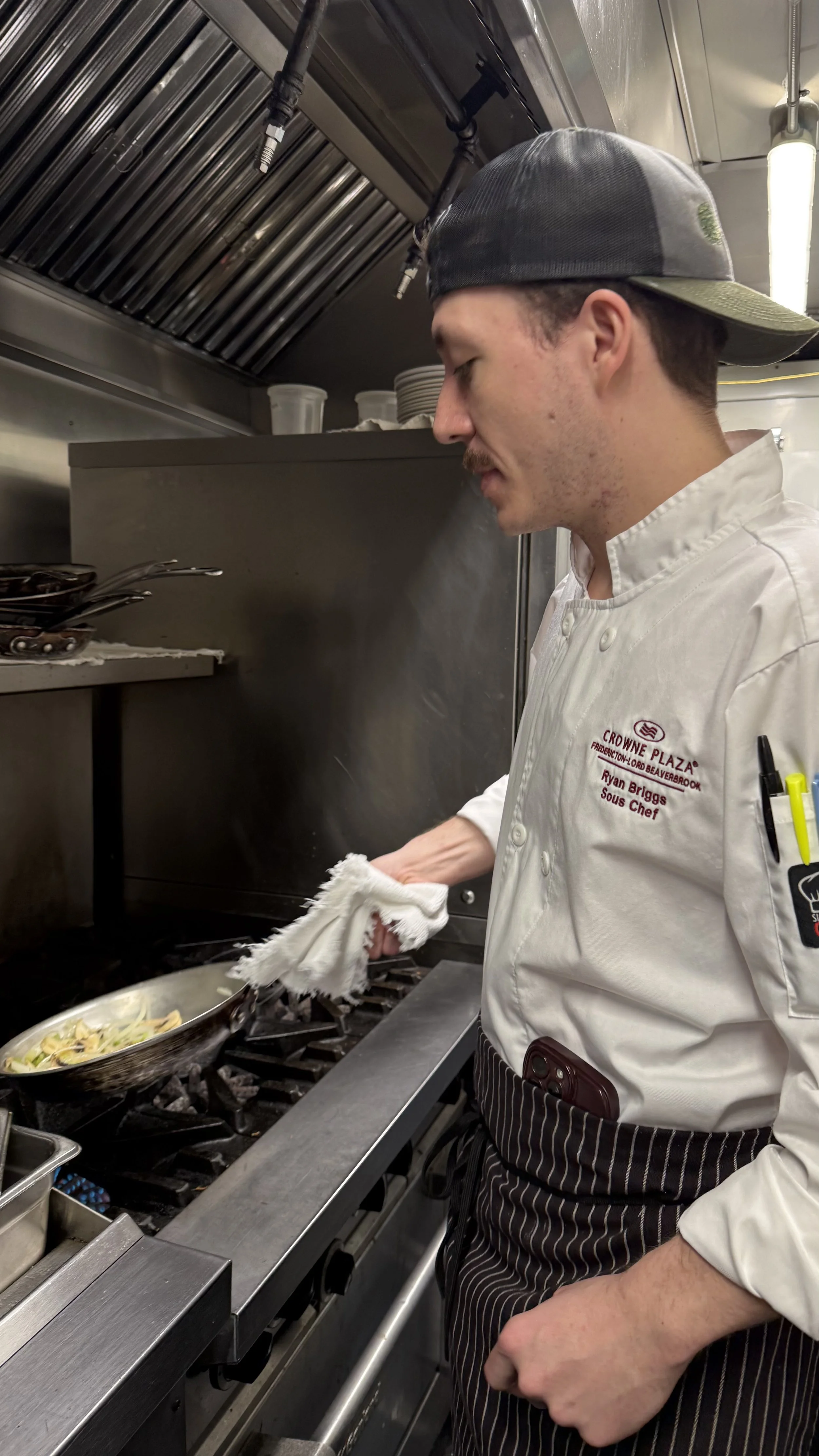 Chef Ryan cooking pasta in Maxwell's kitchen, wearing a white chef's jacket with embroidered text and a black cap, holding a towel near a stove with a pan of pasta.