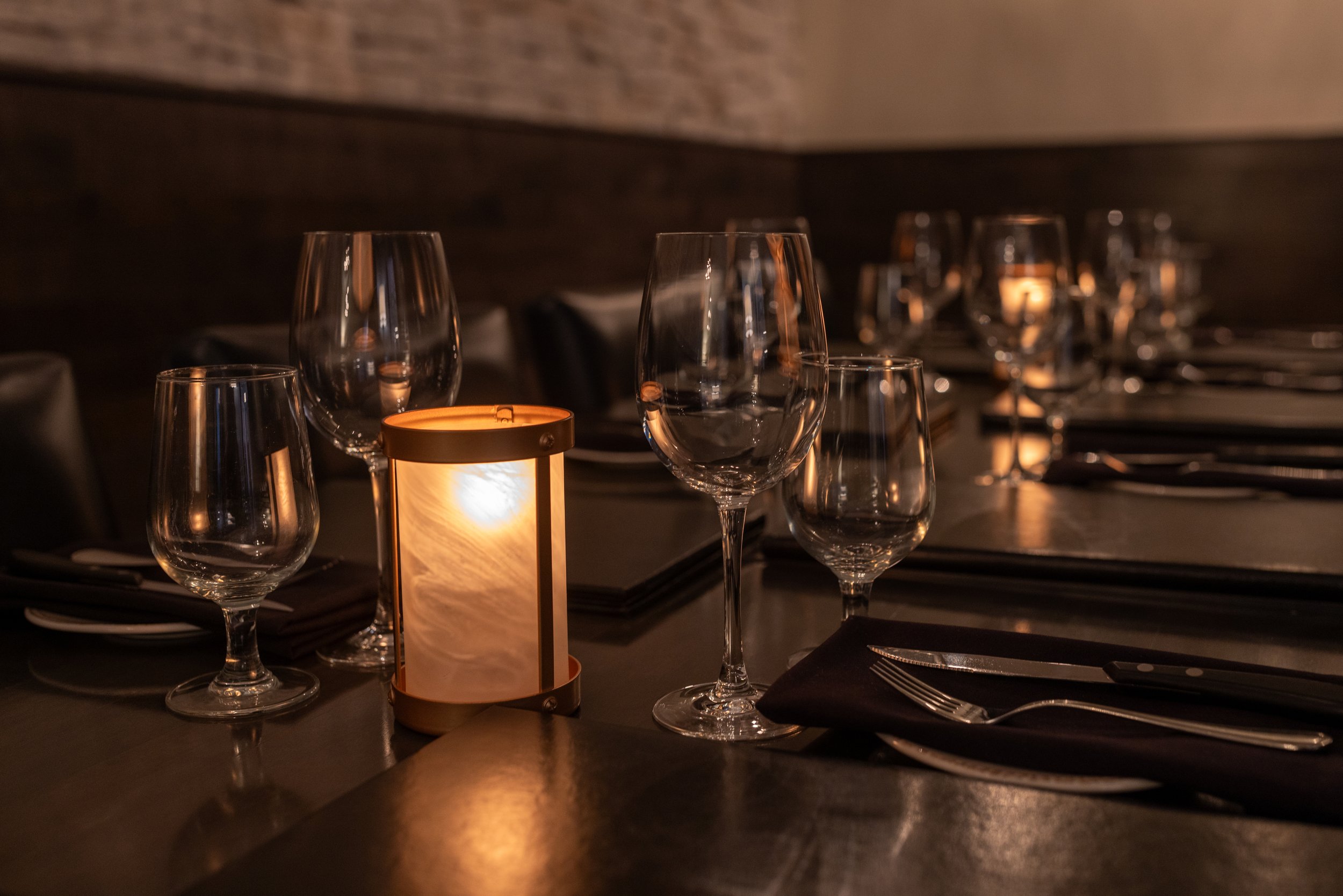 The dimly lit Maverick Room of Maxwell's Steak & Seafood's table set with wine glasses, black napkins, silver cutlery, and a warm candle centerpiece.
