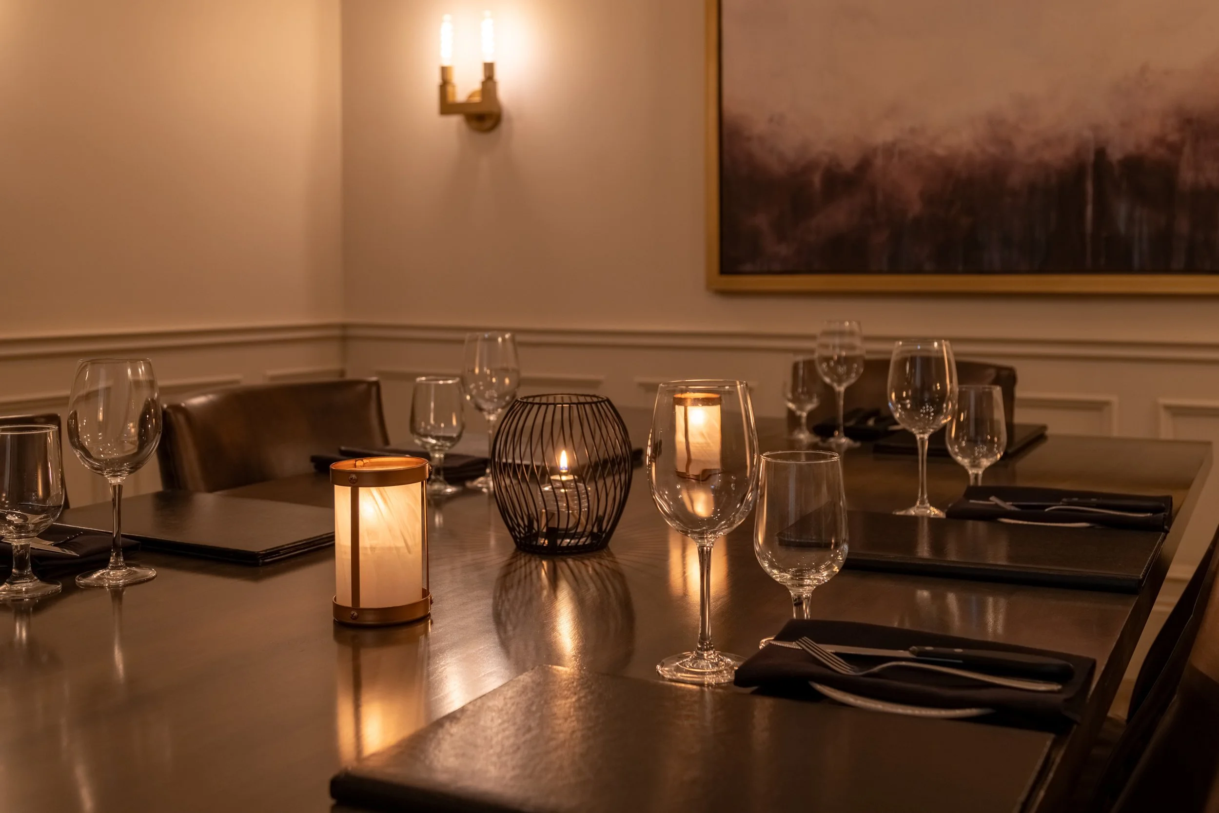 The dimly lit Websters room of Maxwell's Steak & Seafood with a long wooden table set for a meal, featuring wine glasses, black napkins, and candles