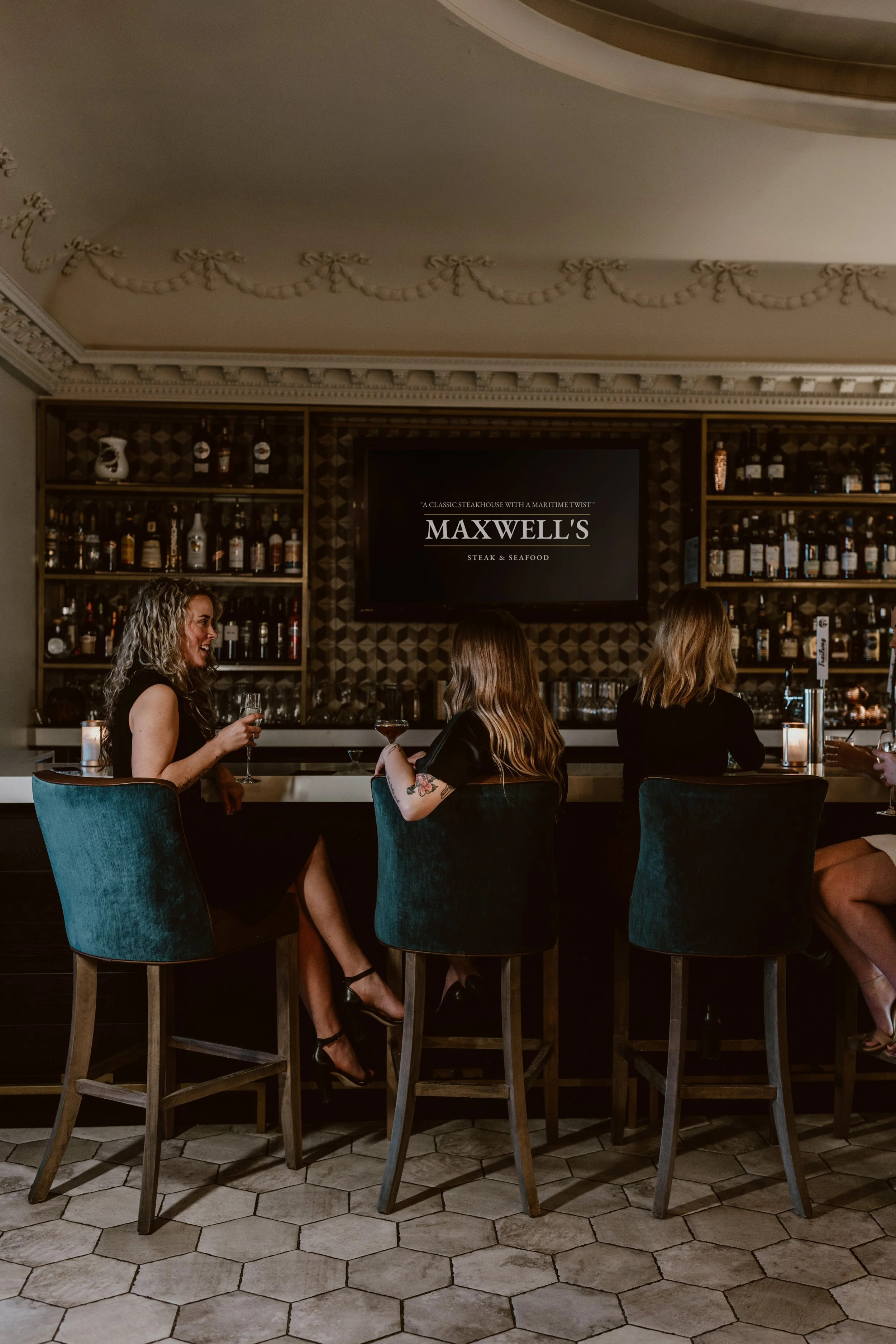 Three women sitting at Maxwell's Steak & Seafood bar, talking and drinking wine in a dimly lit upscale bar or restaurant. Shelves with liquor bottles are visible behind them.