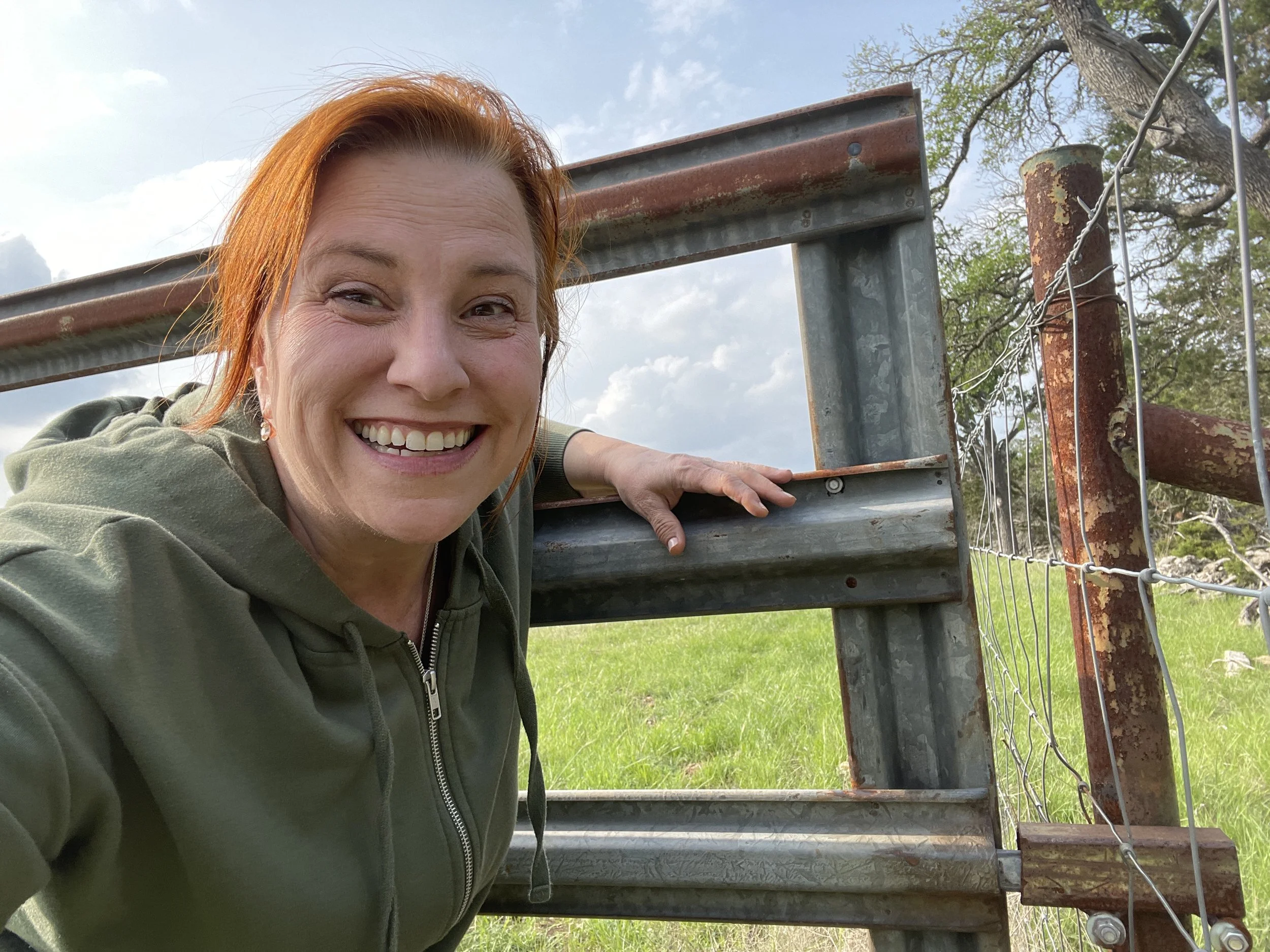 Smiling woman with reddish hair in a green hoodie taking a selfie next to a rusty metal gate and wire fence in an outdoor grassy area, with trees and cloudy sky in the background.