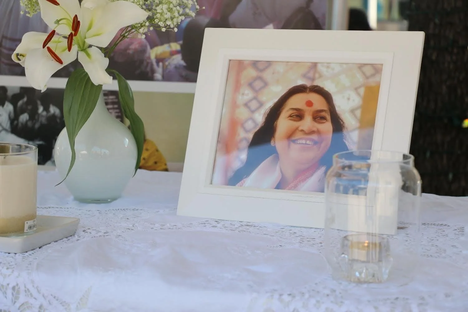 A framed photograph of a smiling woman with a red bindi, placed on a table with a white lace tablecloth. Next to the photo, there are two candles, one in a glass container and another in a small holder, and a white vase holding white lilies and other flowers.