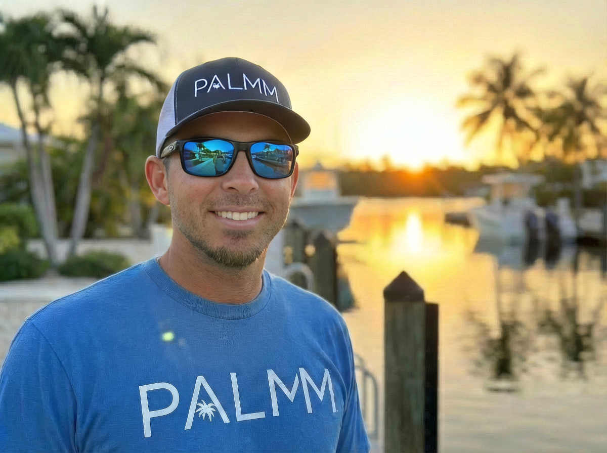Man in blue T-shirt and sunglasses standing at a marina during sunset with boats and palm trees in the background.
