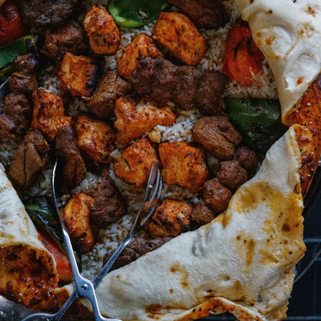 Close-up of a tray full of Mediterranean food with rice, grilled meats, roasted vegetables, and slices of bread or pita.