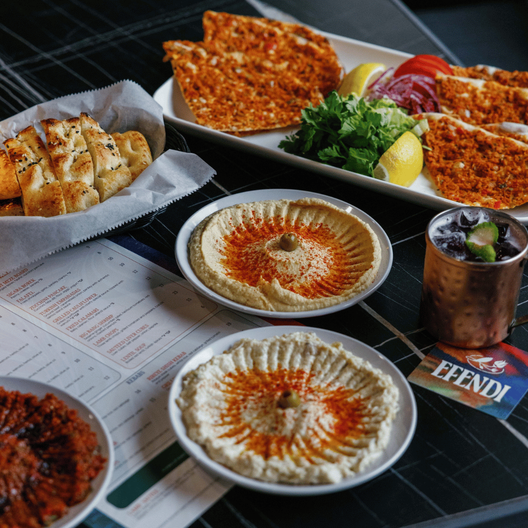 Assorted Middle Eastern dishes on a black table, including hummus, Turkish flatbread with sesame seeds, and Lahmacun topped with vegetables and herbs.