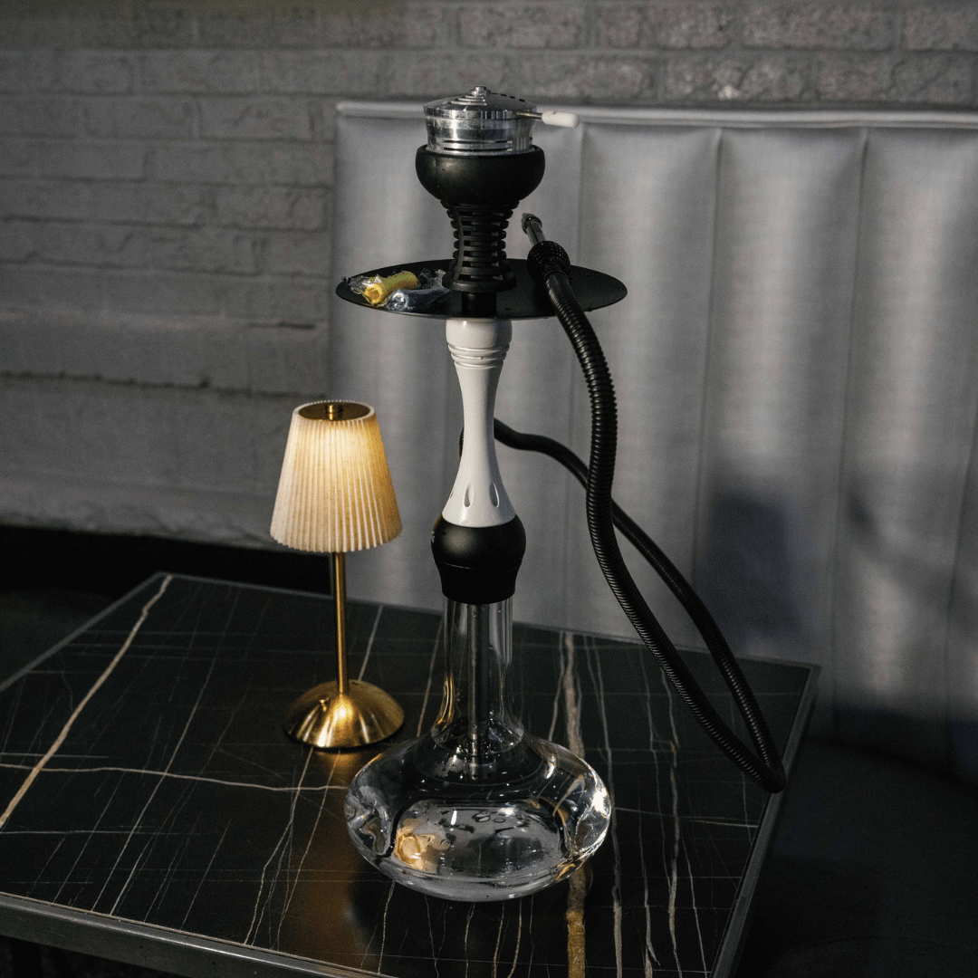 A modern hookah with a glass base, black and white stem, and black hoses, set on a black marble table alongside a small gold desk lamp with a beige lampshade, in a room with a gray brick wall and a white padded headboard.
