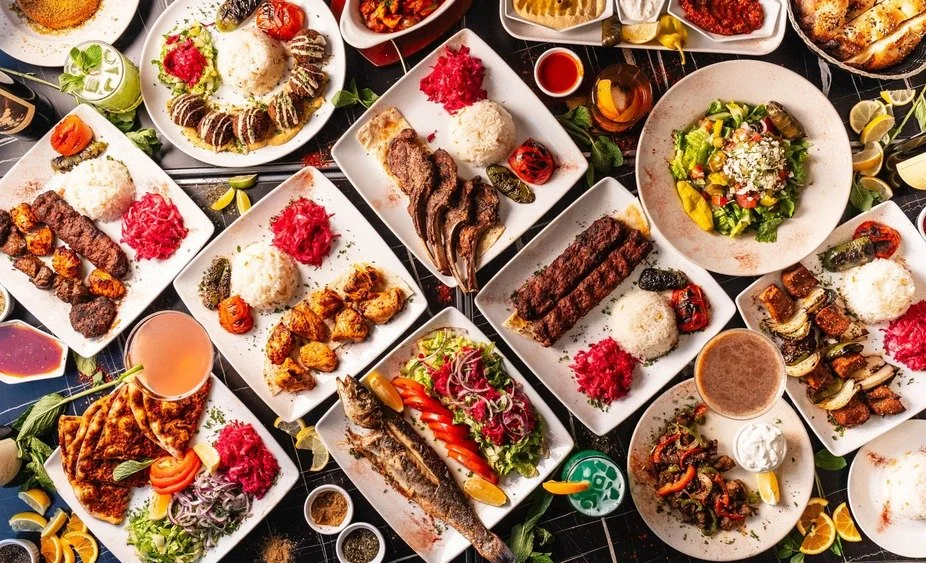 A table filled with various Middle Eastern dishes, including rice, grilled meats, salads, roasted vegetables, and drinks.