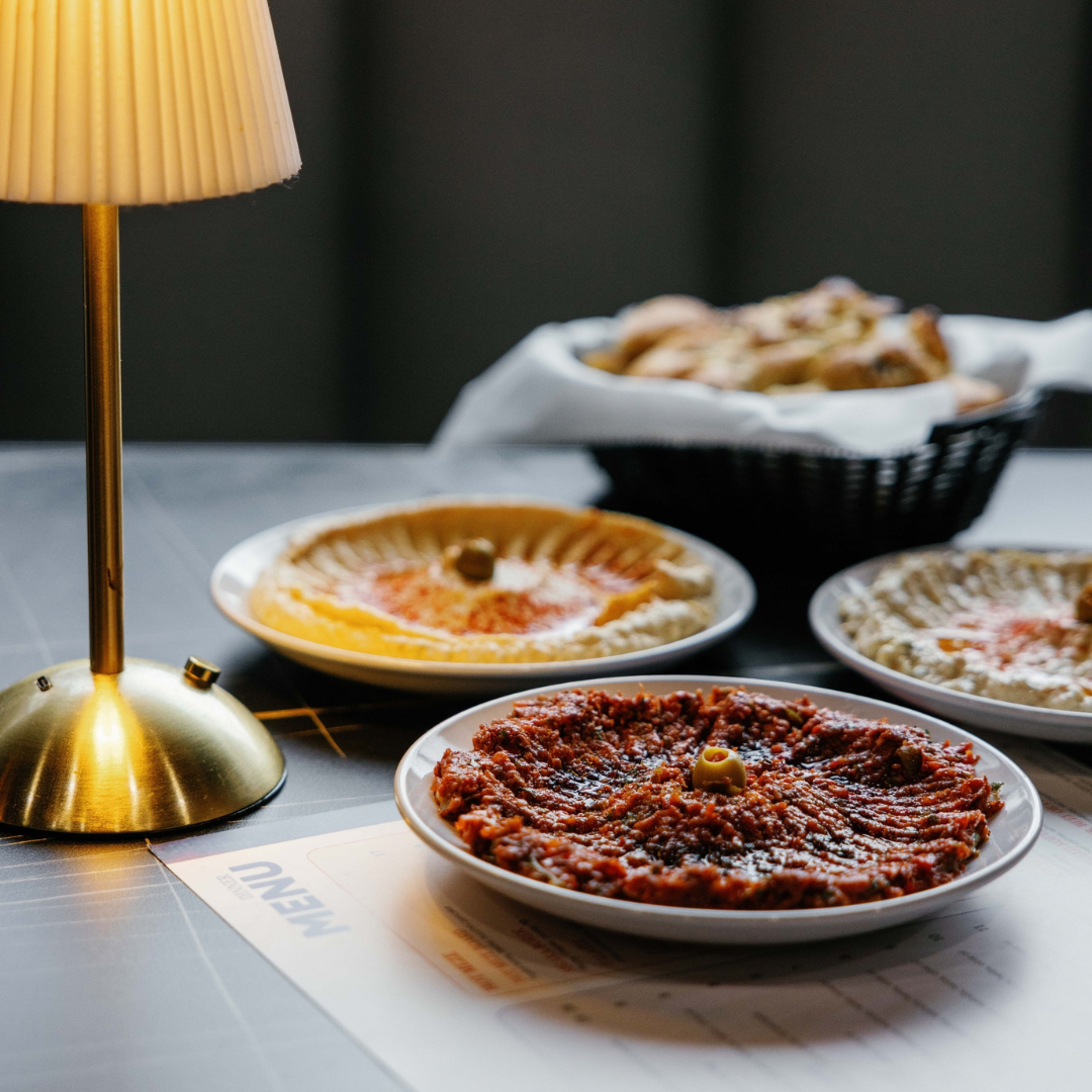 Three plates of different types of pizza on a table near a brass table lamp, with a basket of bread in the background.