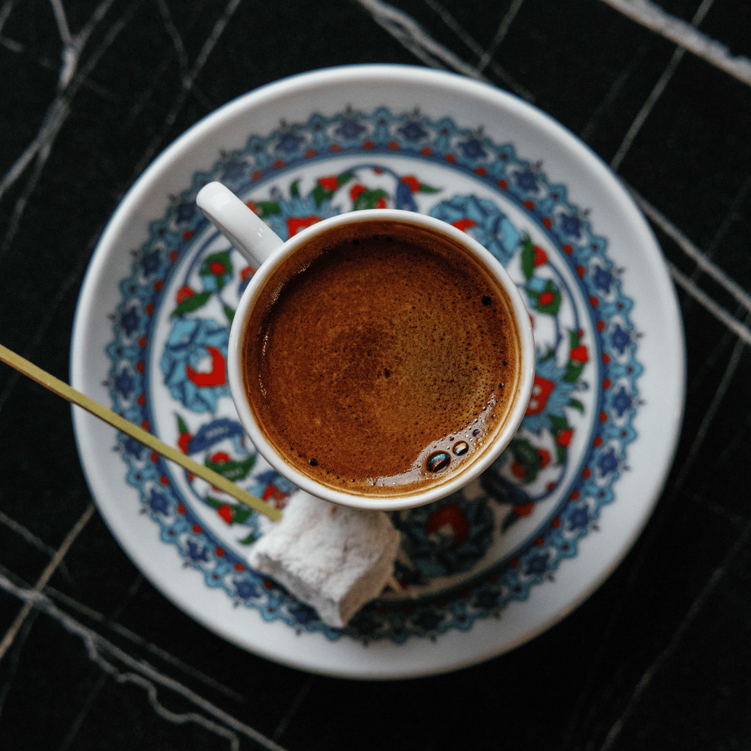Top-down view of a white coffee cup filled with Turkish coffee on a decorative patterned ceramic tray, with a small white sugar cube and a stirring stick.