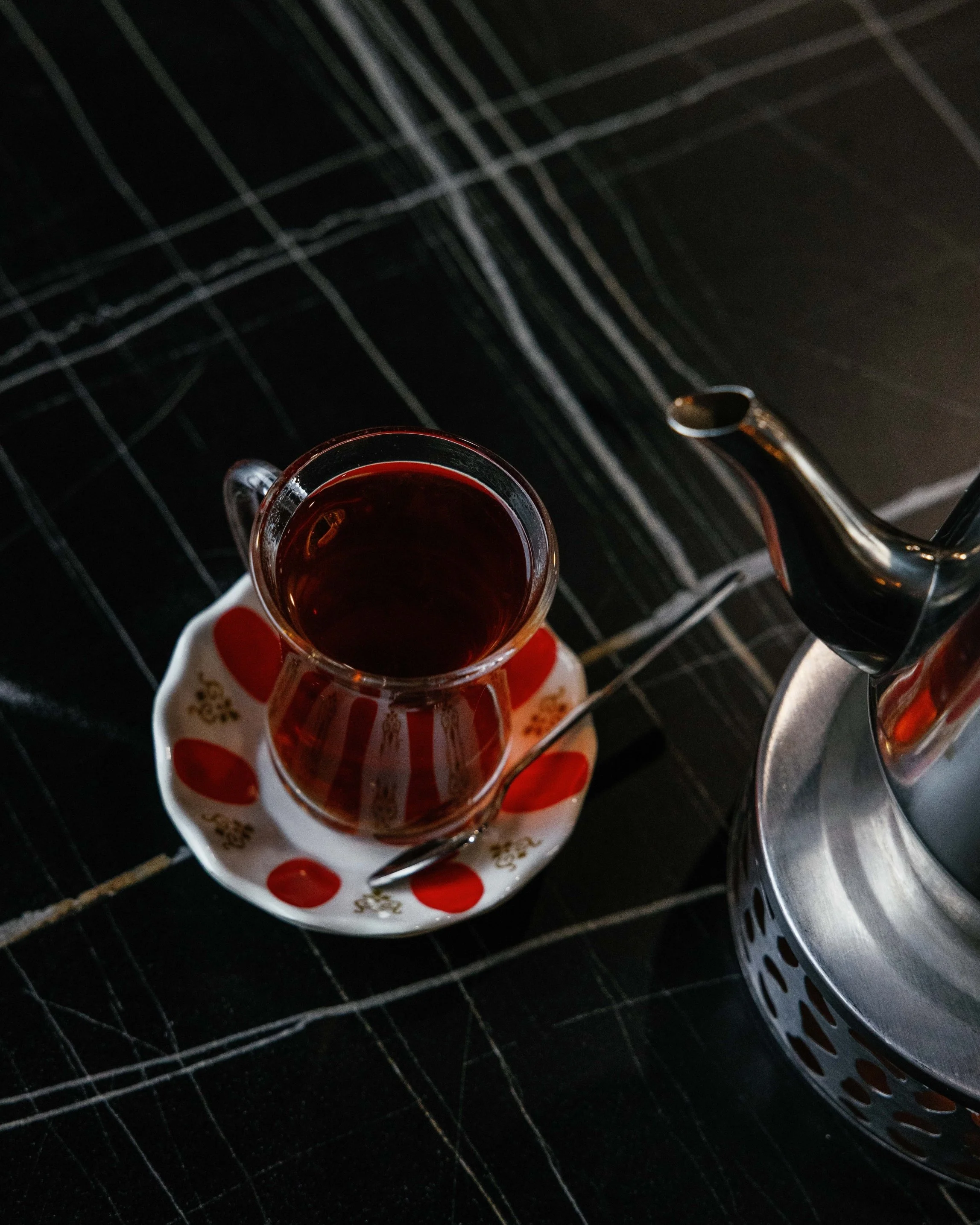 A glass cup of tea on a decorative saucer with red and gold patterns, placed on a table next to a stainless steel teapot.