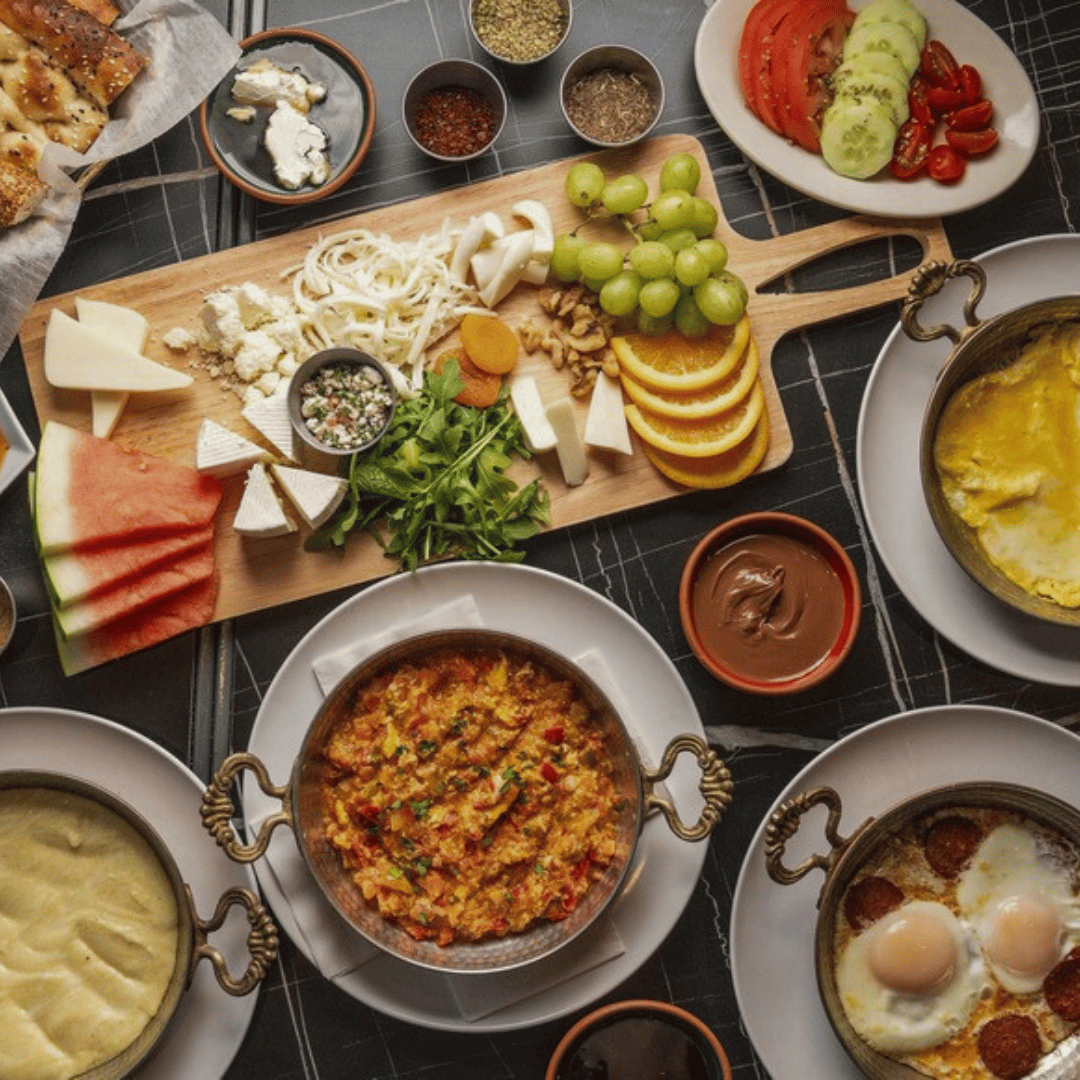 A spread of Middle Eastern breakfast foods including scrambled eggs, eggs cooked in tomato sauce with meatballs, hummus, bread, a cheese platter, watermelon slices, fresh vegetables, grapes, and various spices and condiments.