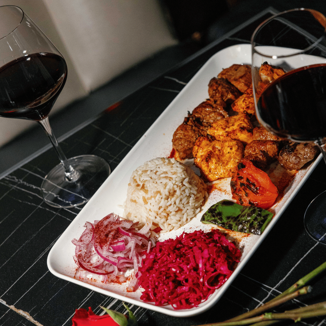 A rectangular white plate with grilled meat, rice, and colorful salads, accompanied by two glasses of red wine, on a black table.