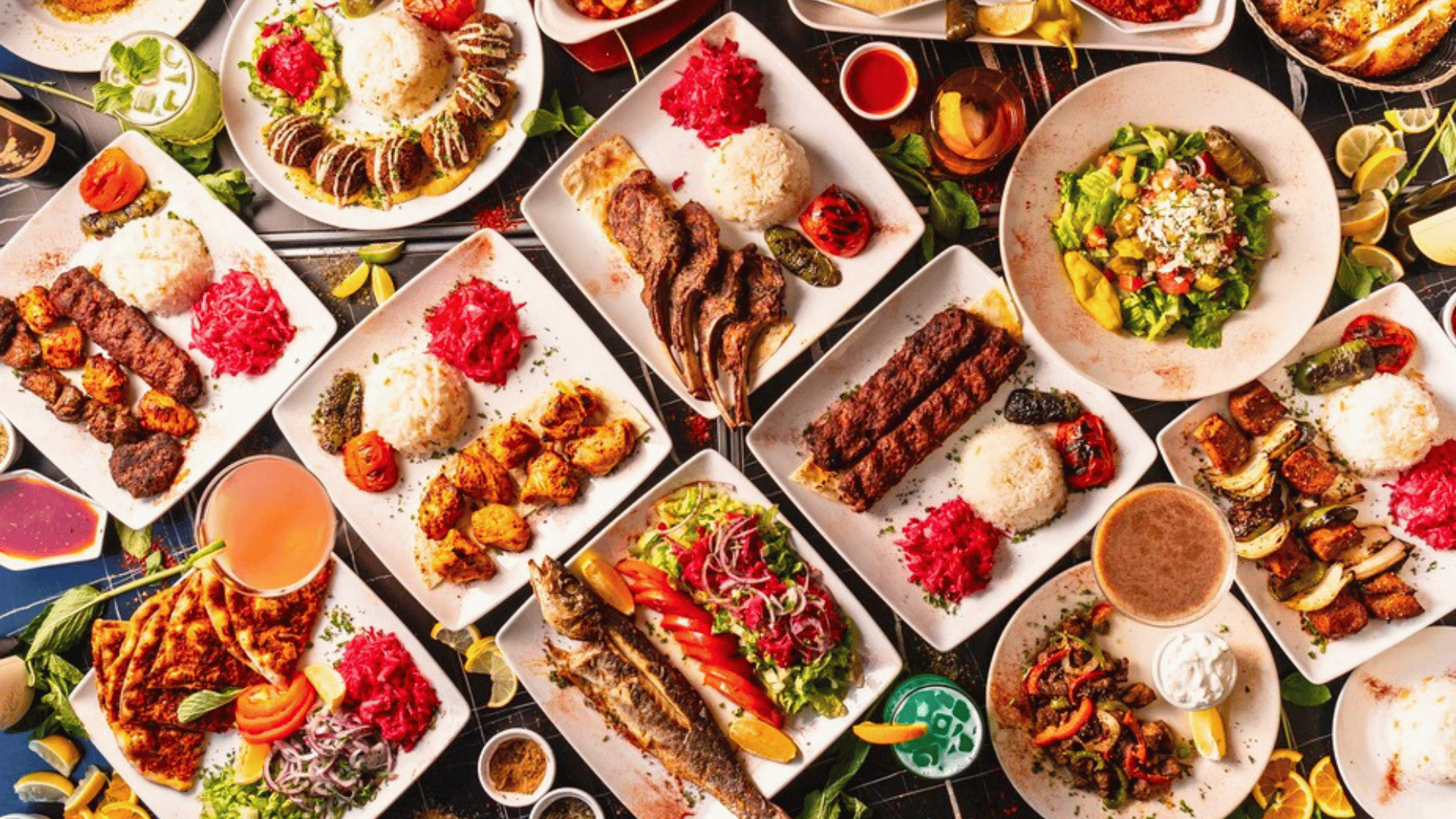 A variety of Middle Eastern dishes on plates and bowls, including grilled meats, rice, salads, vegetables, and sauces, with lemon wedges and fresh herbs on a black table.
