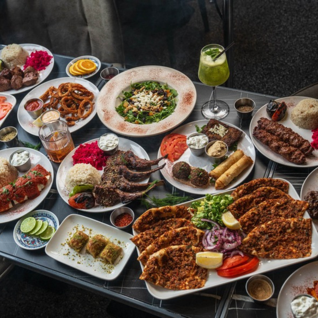 A table filled with various Middle Eastern and Mediterranean dishes including grilled meats, salads, rice, and beverages, served on white plates and bowls.