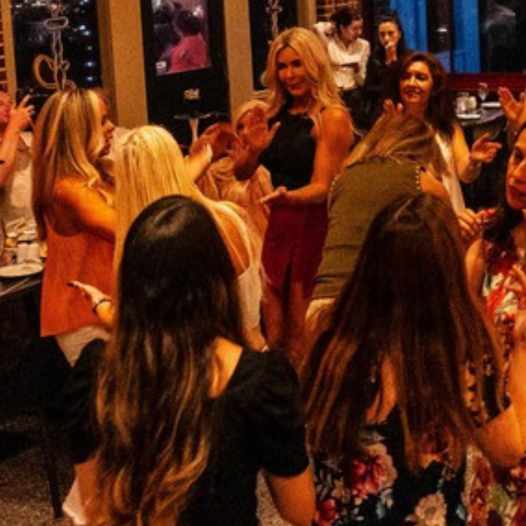 A group of women at a social gathering or party, some standing and some sitting, engaging in conversation and socializing in a warmly lit indoor setting.