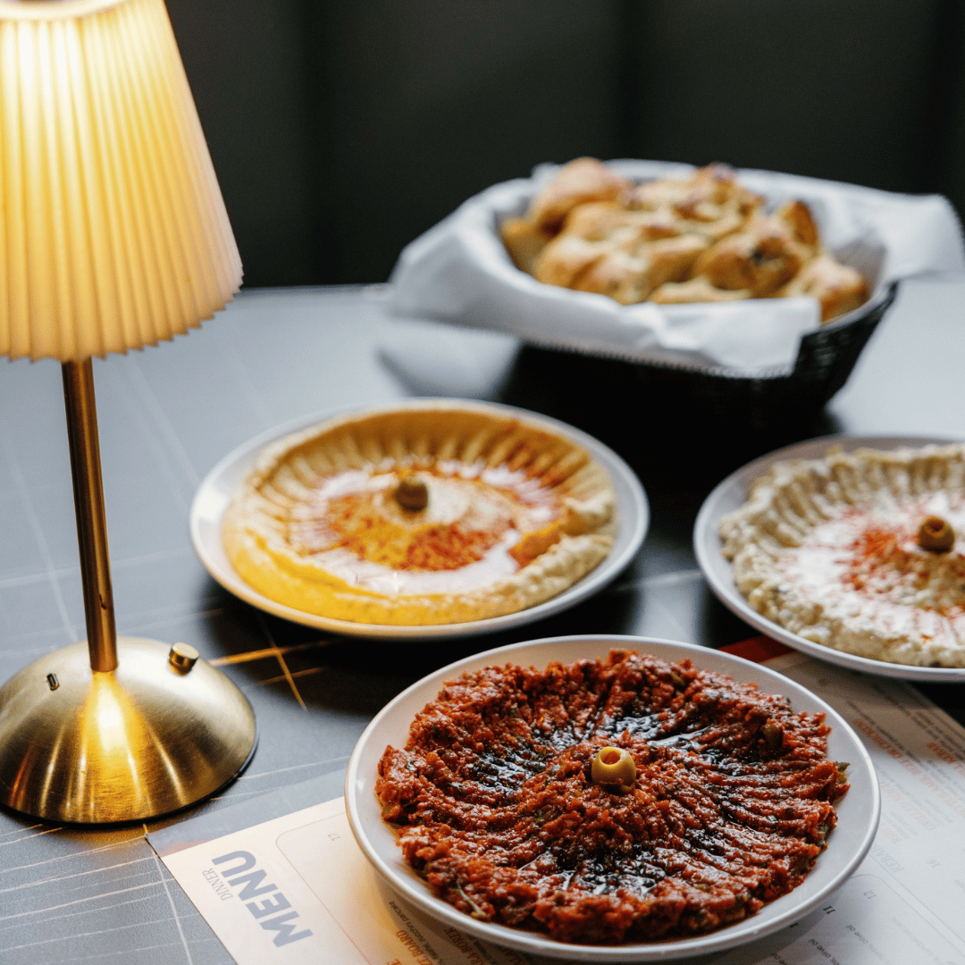 Four plates of Italian antipasti on a table, with a lamp and a basket of bread in the background.