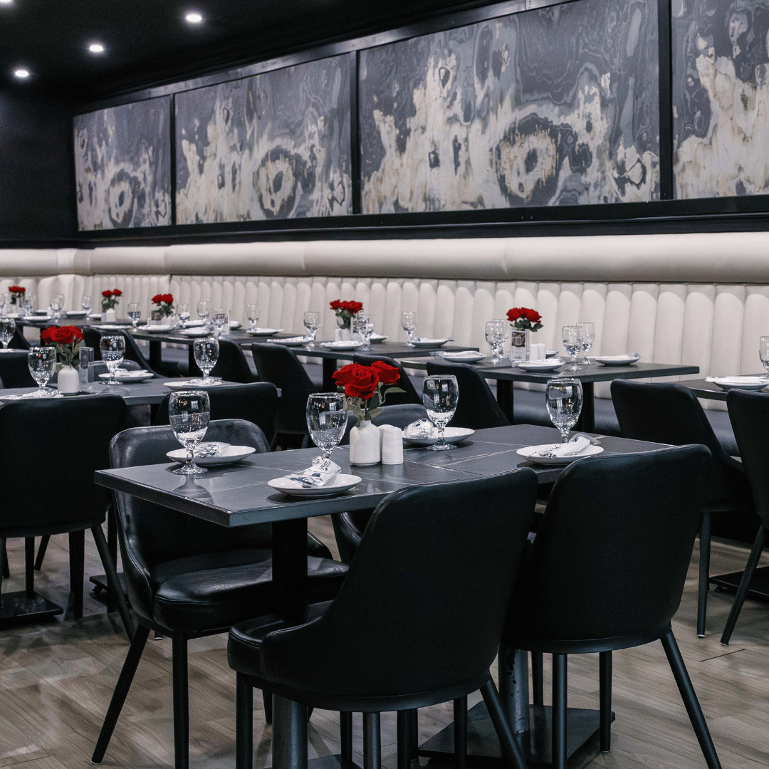 Elegant restaurant with black tables, black chairs, and white upholstered wall padding. Each table has a red rose centerpiece, water glasses, and white plates with napkins.