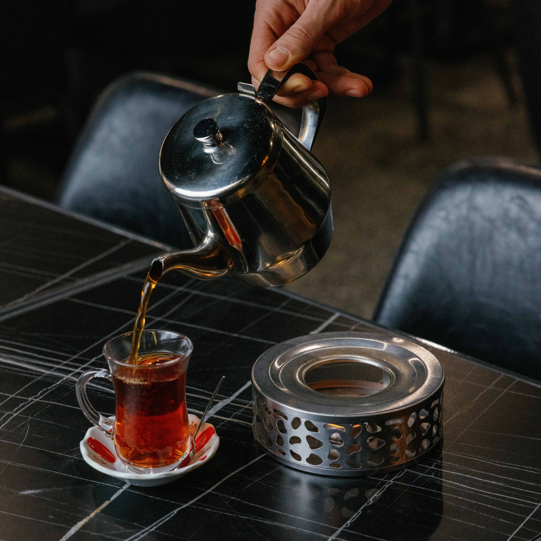 A hand pouring hot tea from a stainless steel kettle into a glass teacup on a small saucer on a black table.