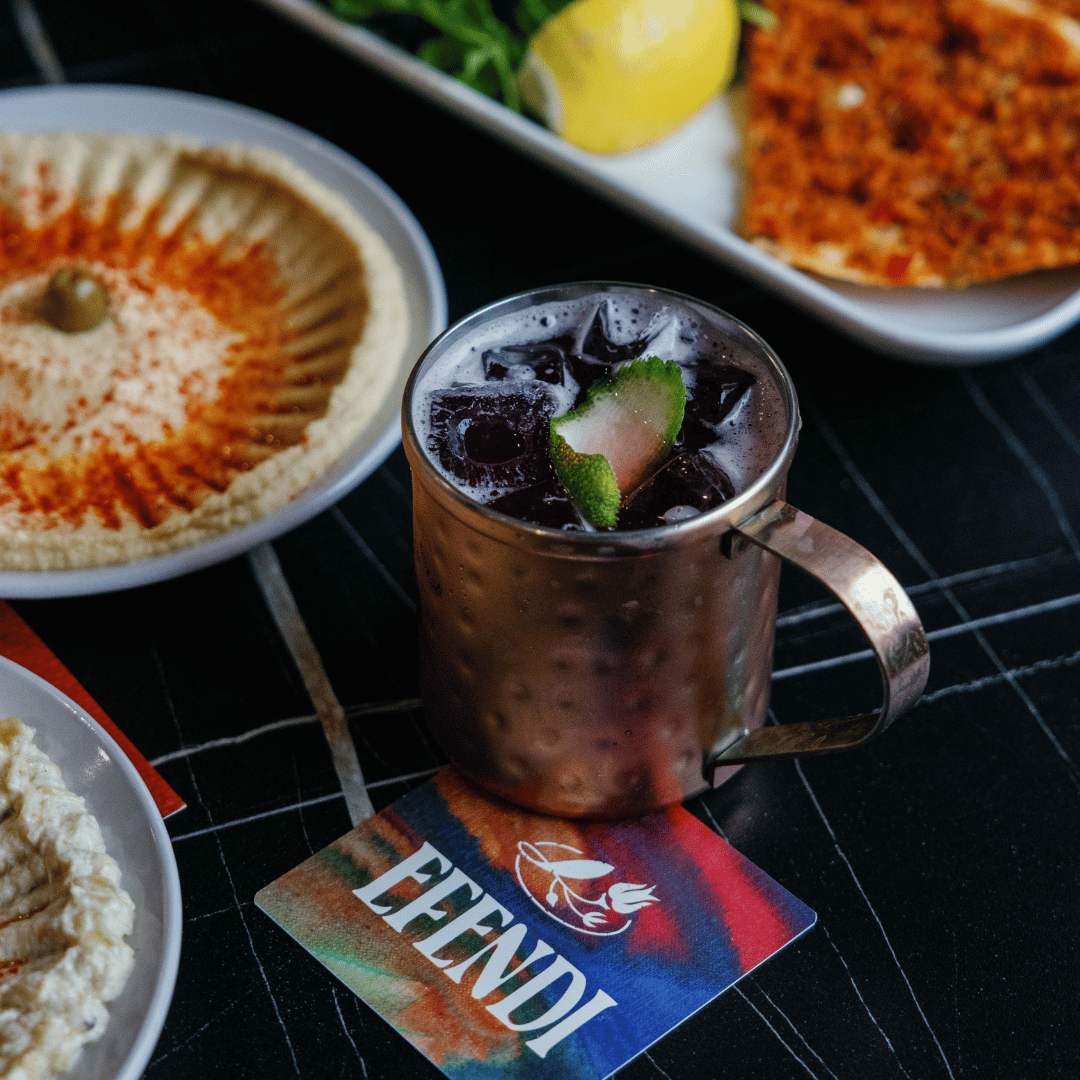 A glass of iced beverage with ice cubes and lime slice on a colorful coaster, surrounded by plates of hummus with paprika, a slice of flatbread, and another dish with a yellow lemon and possibly more flatbread, on a black surface.
