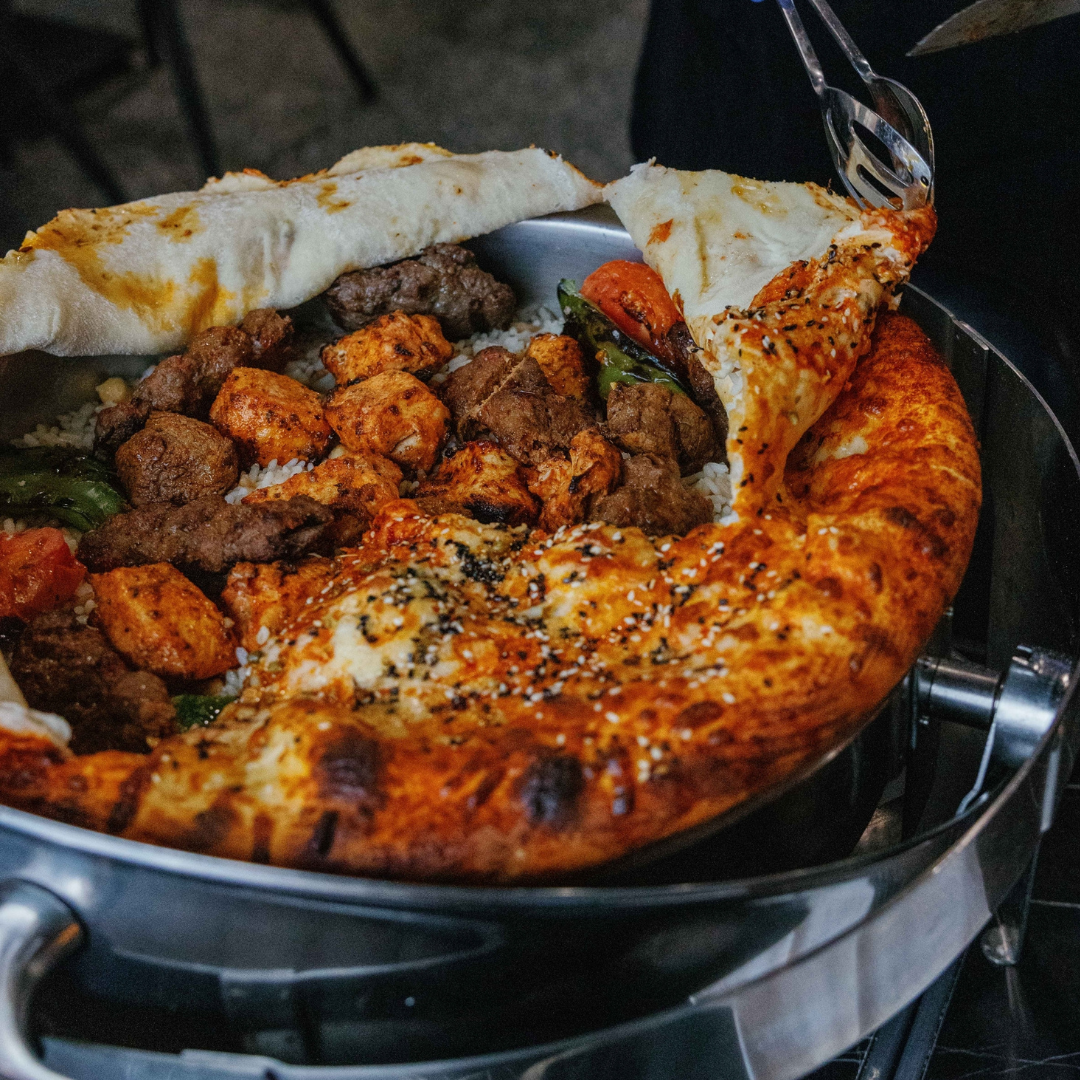 A close-up of a hot Mediterranean or Middle Eastern dish with pide bread, rice, grilled meat, and vegetables, topped with sesame seeds.