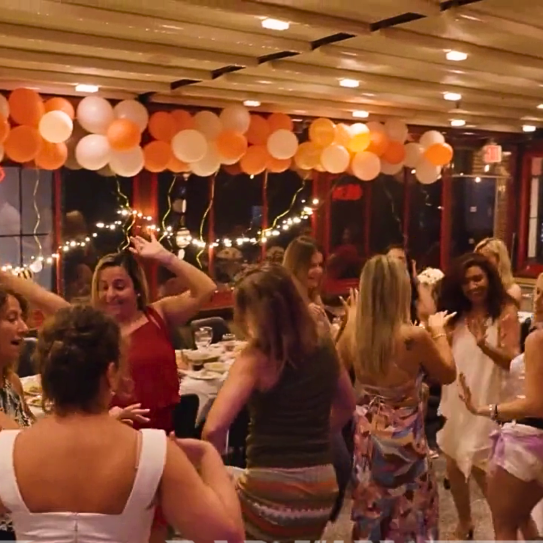 A group of women dancing and celebrating at a party with orange and white balloons and string lights in the background.