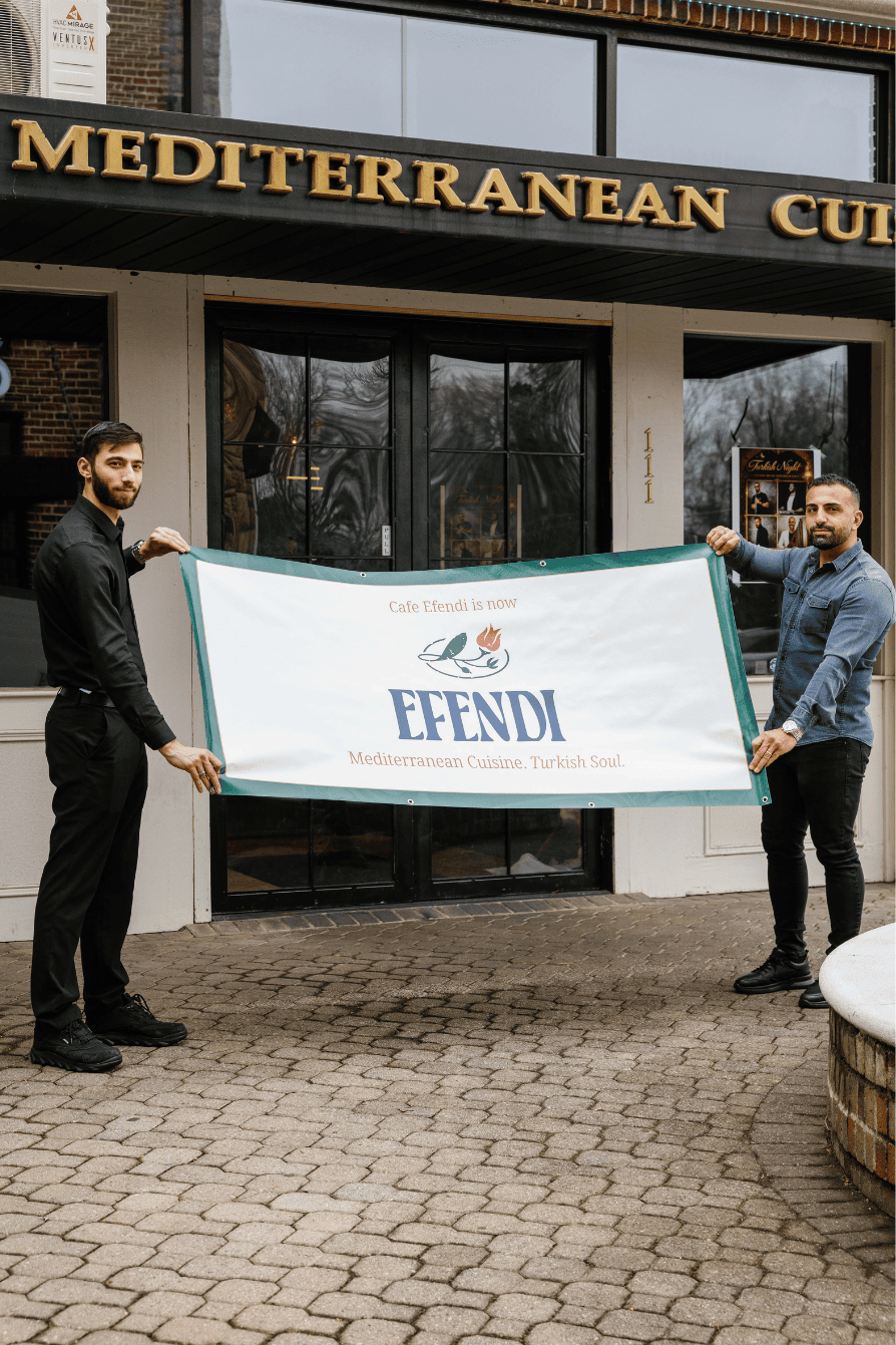 Two men holding a banner outside a Mediterranean Cuisine restaurant called EFENDI, with the sign 'Cafe Efendi is now EFENDI Mediterranean Cuisine. Turkish Soul.'
