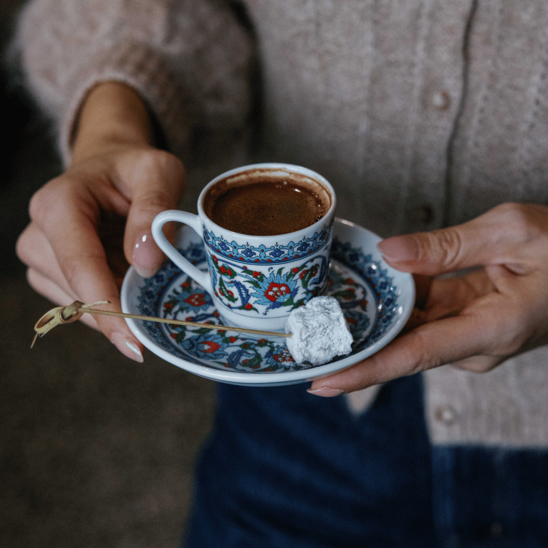 Person holding a decorative blue and white tray with a small cup of Turkish coffee and a piece of Turkish delight covered in powdered sugar.