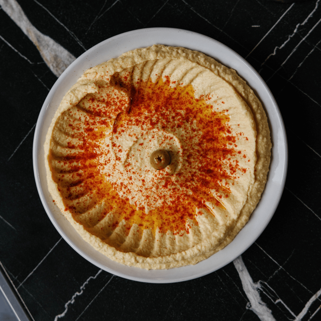 A bowl of hummus garnished with paprika and an olive on top, placed on a black marble surface.