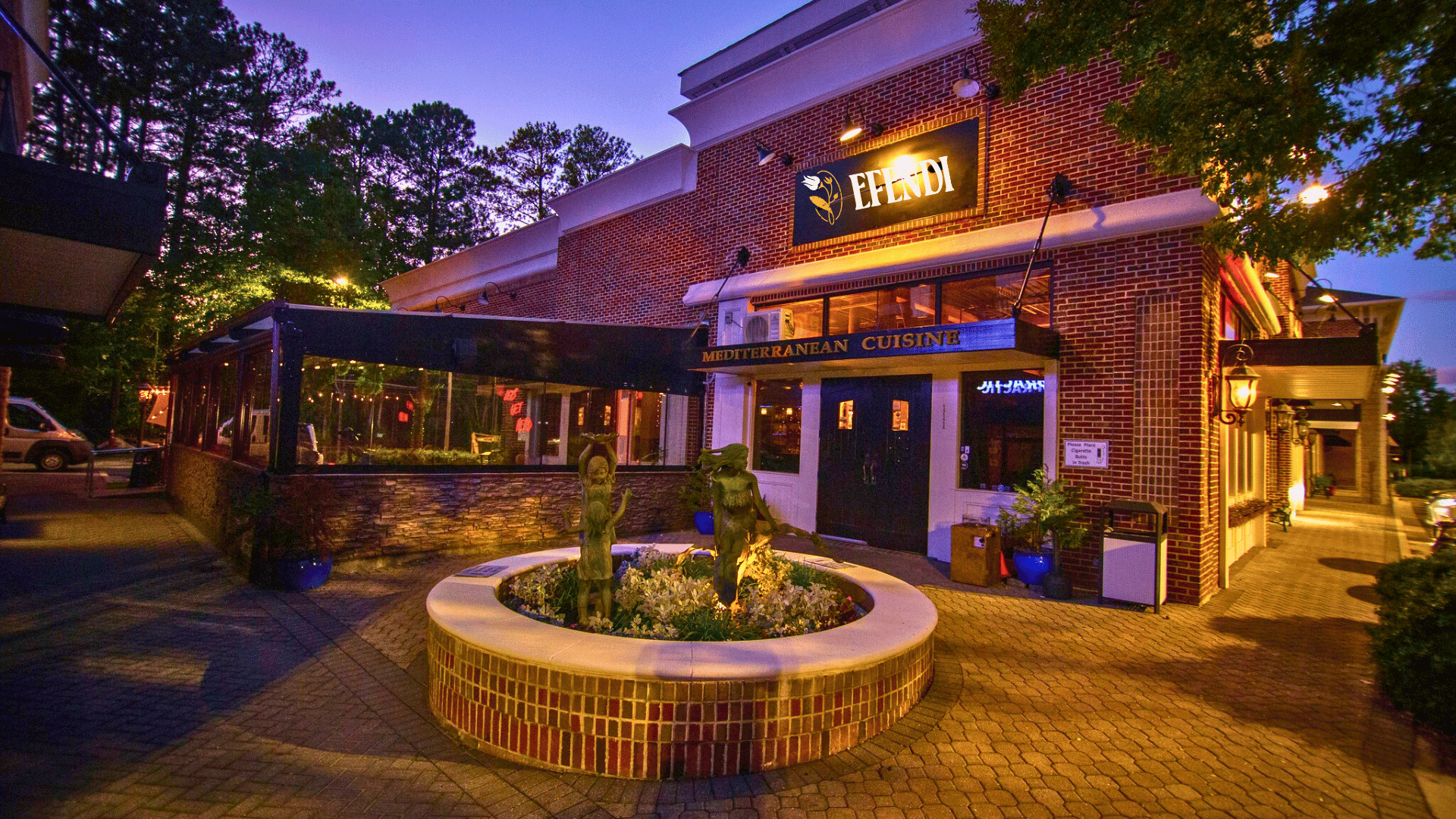 An exterior view of a Mediterranean Cuisine restaurant named Efendi, with a brick facade, outdoor seating area, and a small fountain with sculptures in the foreground during evening hours.