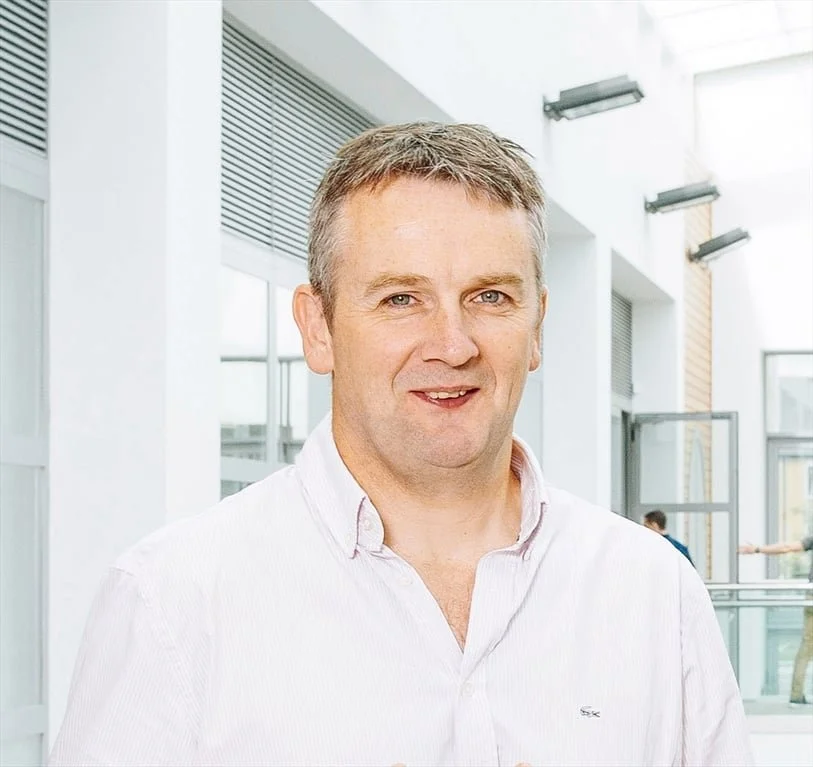 A middle-aged man with short, light brown hair, wearing a white collared shirt, standing inside a modern building with large windows and natural lighting.