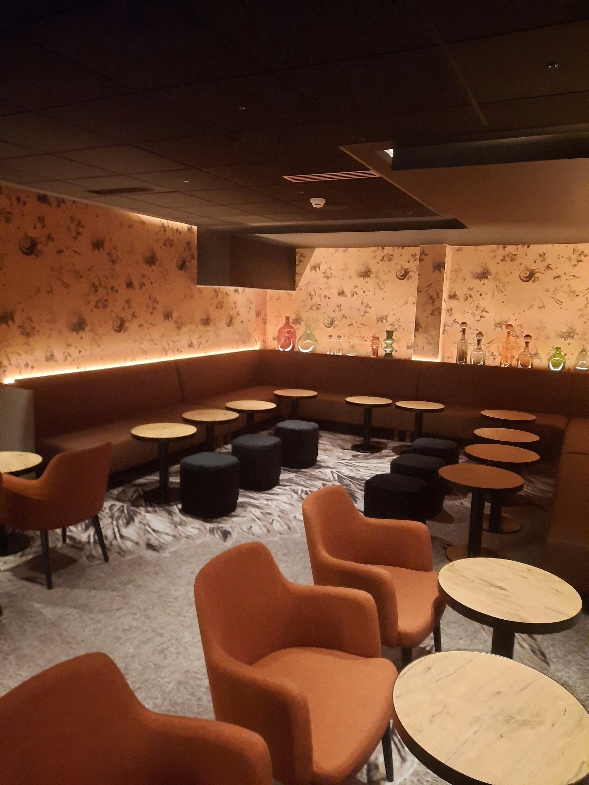 Un salon de lounge élégant avec des chaises orange, des tables de marbre et un mur décoratif avec des flacons colorés sur une étagère.