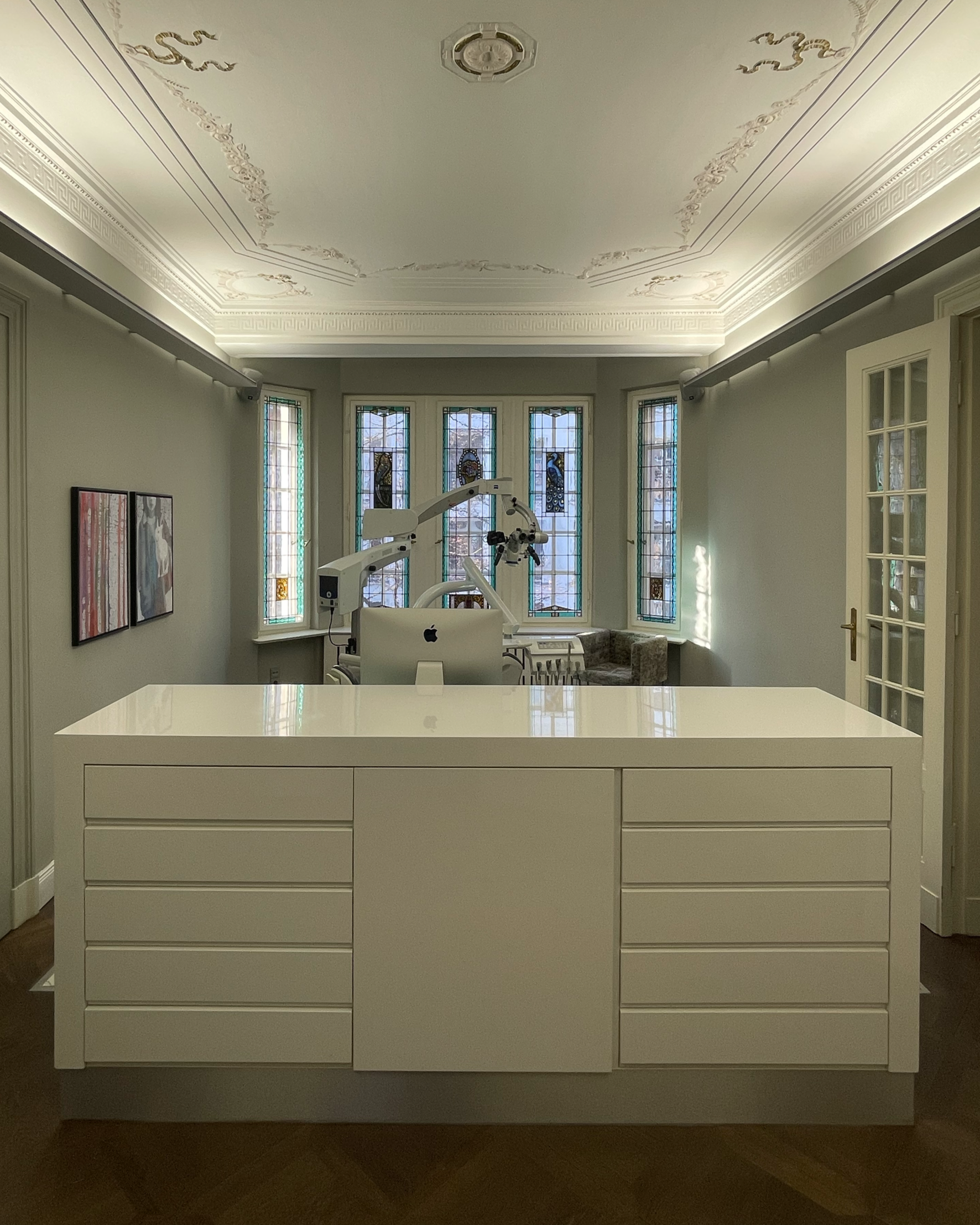 Ein modern eingerichteter Behandlungsraum mit weißen Möbeln, einem Computer und einem Operationsmikroskop vor einem Fenster mit buntem Glas.