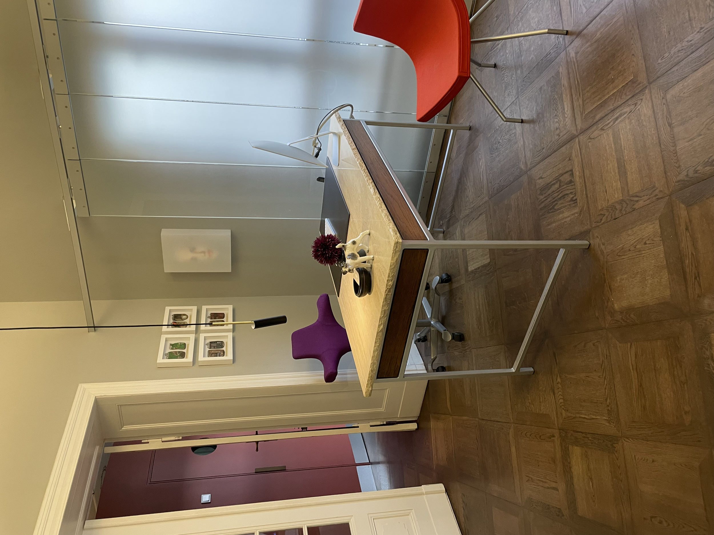 Ein Büro mit einem Schreibtisch, einem roten und einem lila Stuhl, einer Schreibtischlampe, einer Pflanze auf dem Tisch, einem weißen Bilderrahmen an der Wand und einem grauen Schrank in Hintergrund.