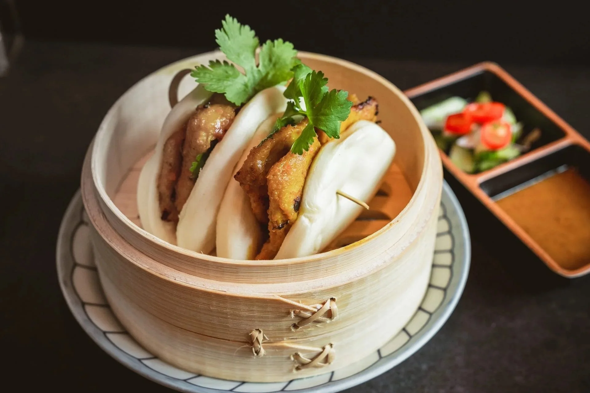 Two steamed bao buns filled with braised meat and garnished with cilantro, served in a bamboo steamer with a side of dipping sauce.