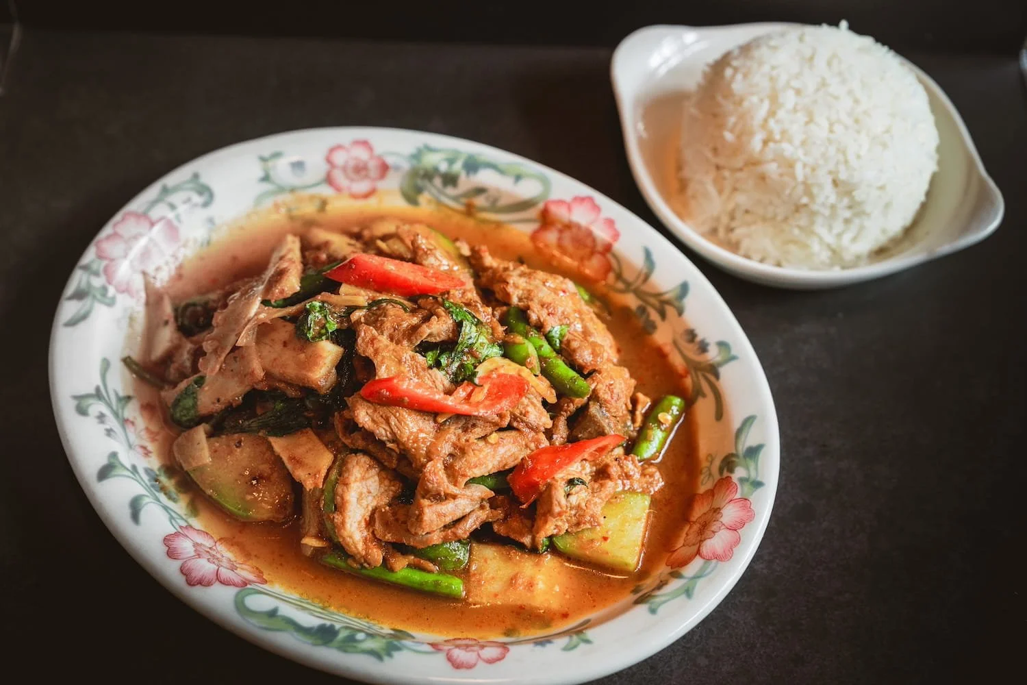 Wok Curry Paste with sliced meat, string beans, red chili, and basil in a rich curry sauce on a floral ceramic plate, served with steamed rice.