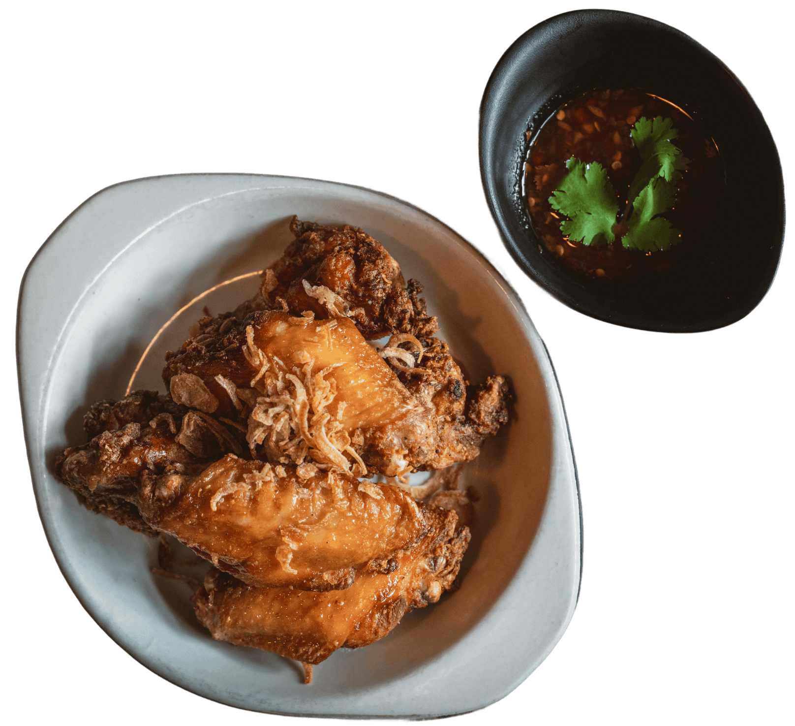 Bottom left is fried chicken wings with crispy golden-brown skin, served on a white oval plate and top right is a small black bowl of dark chili dipping sauce garnished with fresh cilantro.