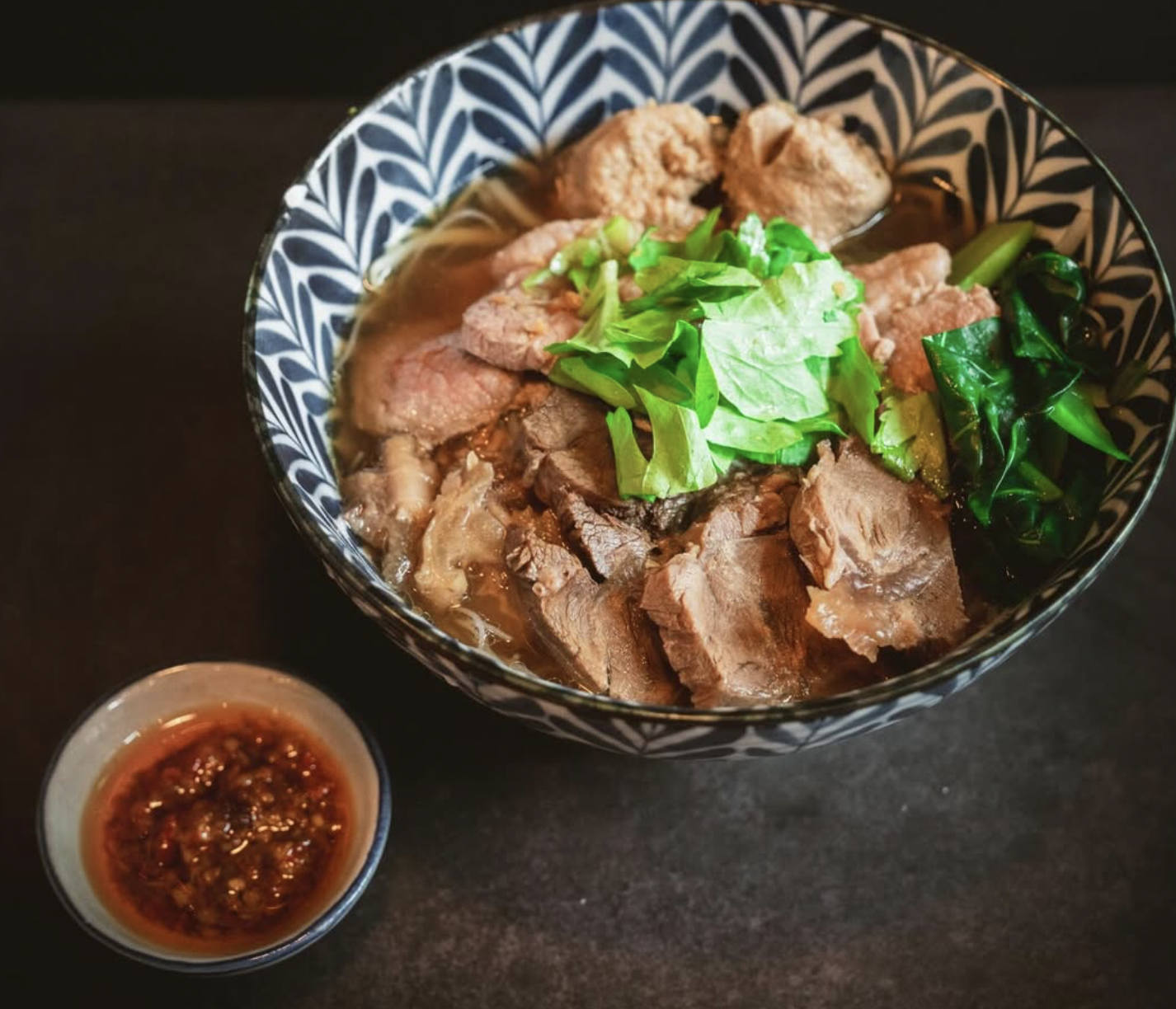 Blue and white patterned bowl of pork noodle soup with thick-cut braised pork, greens, and a side of chili dipping sauce.