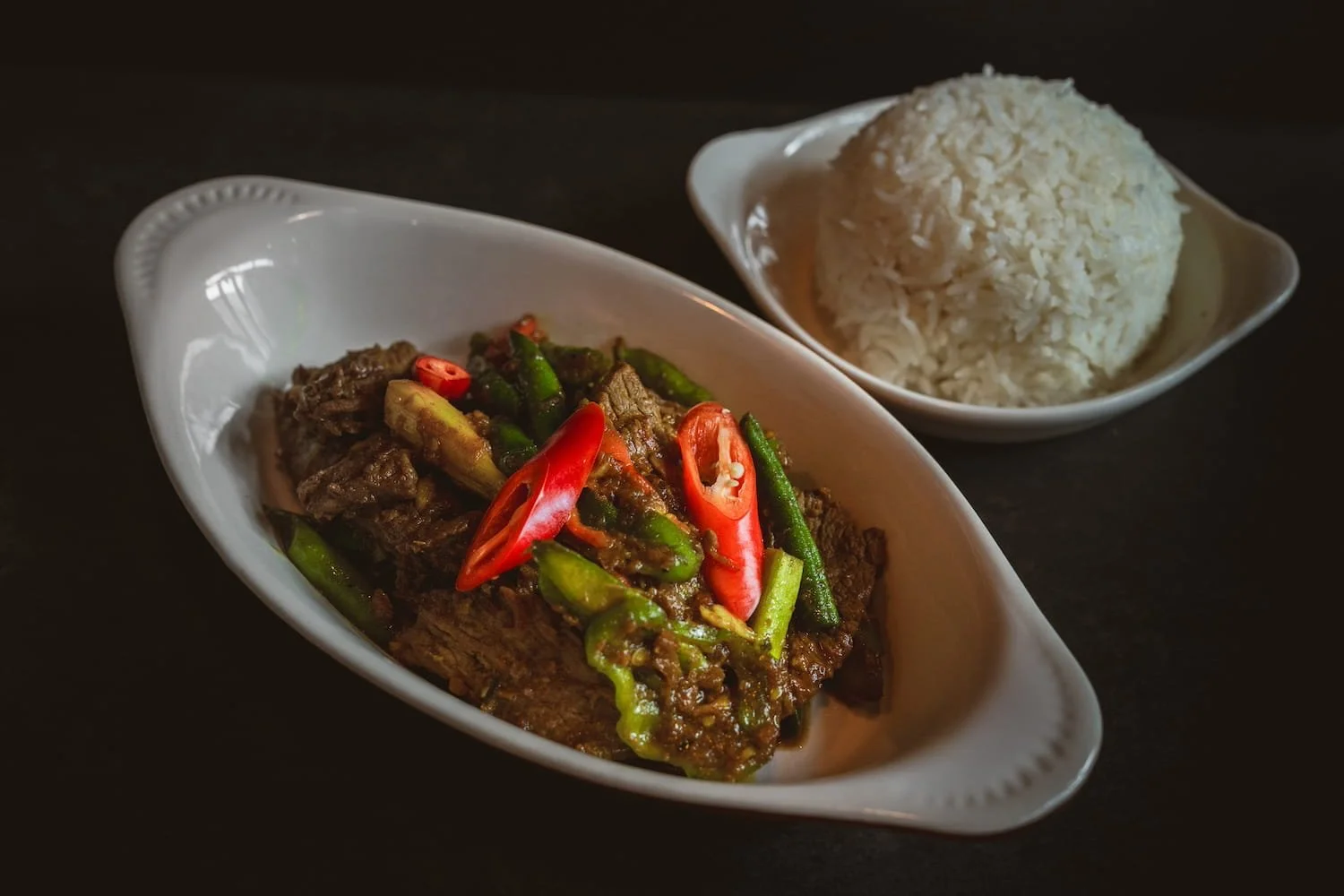 Stir-fried beef with red chili and string beans in a dark sauce in a white oval dish, served with steamed rice.