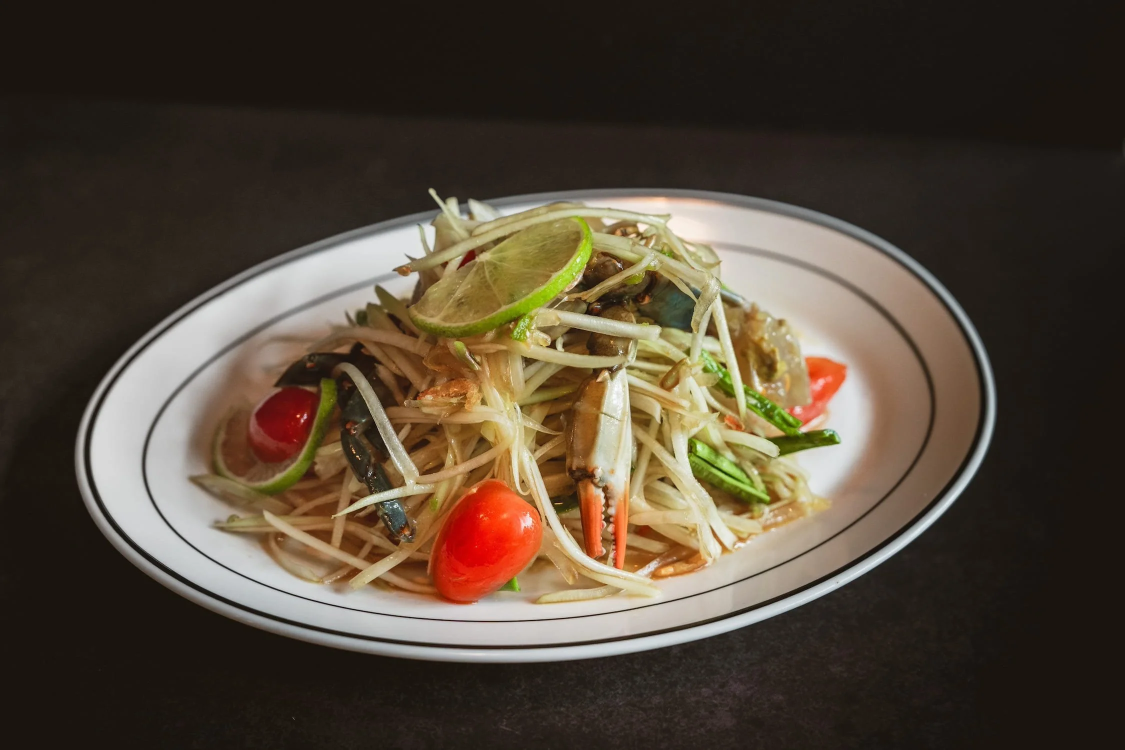 Blue Crab Papaya Salad with shredded green papaya, cherry tomatoes, long beans, and a wedge of lime on an oval white plate.