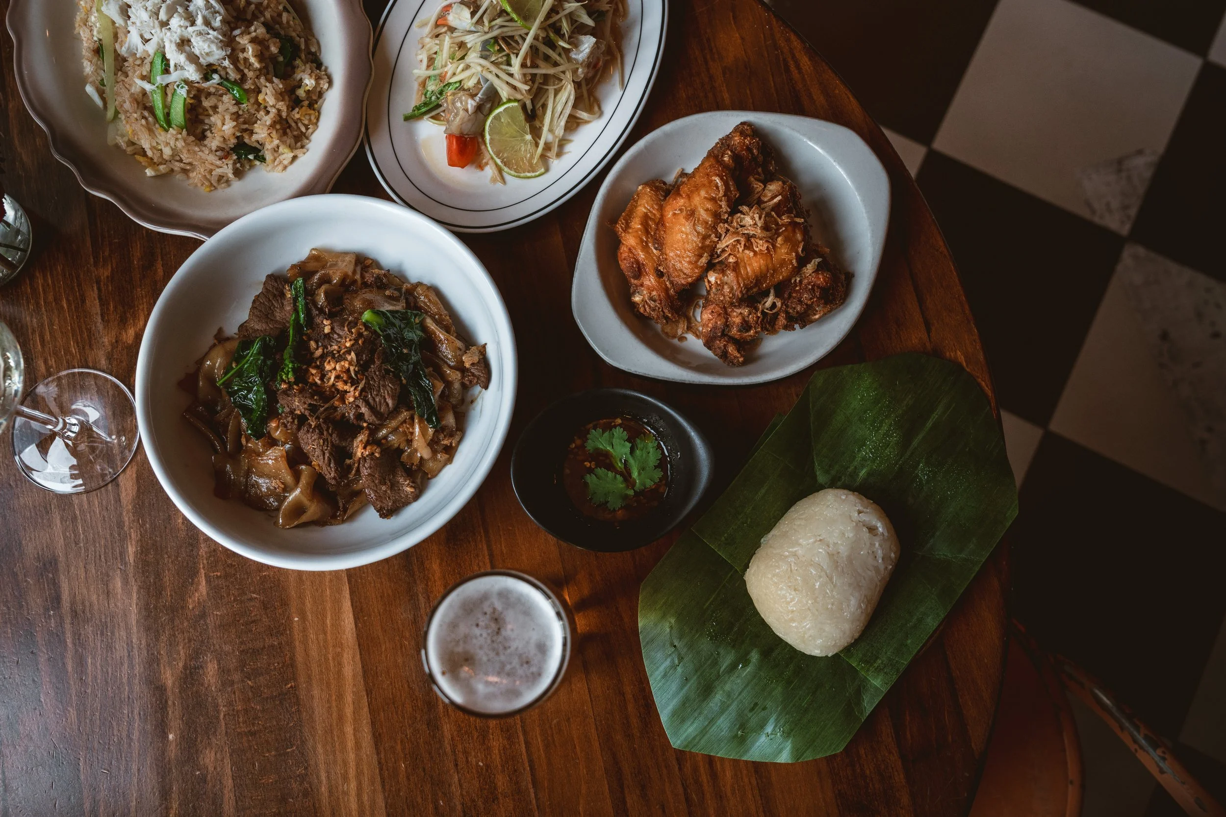 Overhead flatlay of Thai dishes on a wooden table: green papaya salad, fried chicken wings, stir-fried beef with noodles, dark dipping sauce with cilantro, sticky rice on a banana leaf, and a glass of beer.