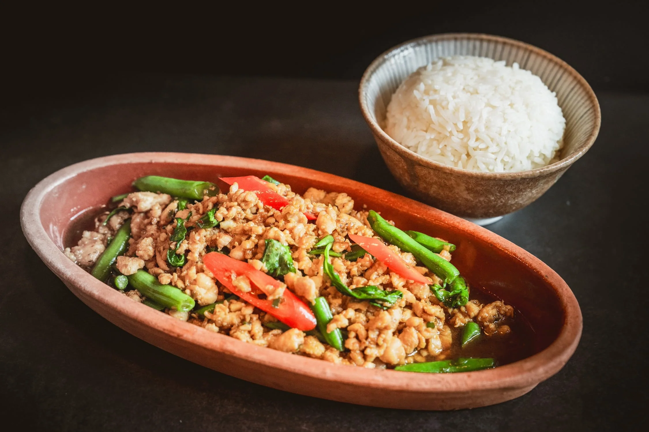 Wok Basil with ground meat, string beans, and red chili in a clay oval dish, served with a bowl of steamed rice.